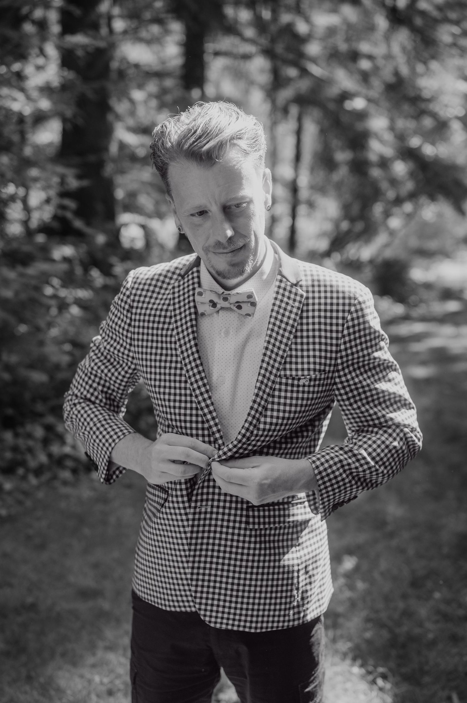 Black and white photo of a stylish man in a checkered blazer and bow tie buttoning his jacket outdoors.