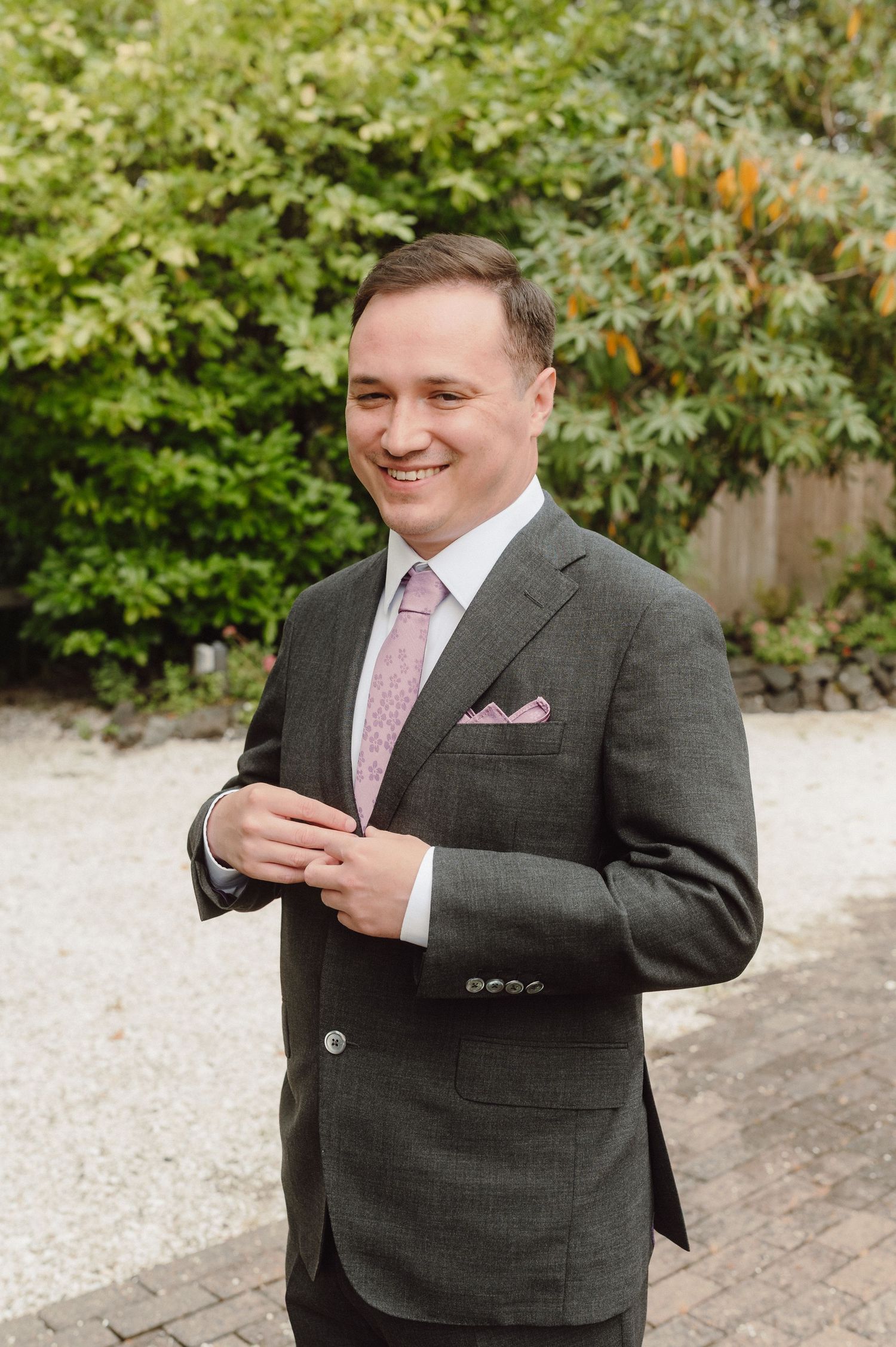 Someone in a grey suit jacket and pink tie smiles while looking at their phone outdoors with greenery in the background.