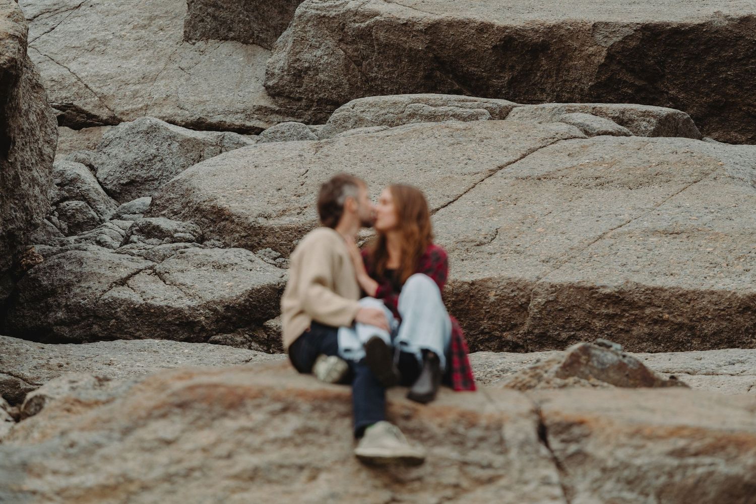 A couple sits together on large rocky outcrops overlooking a scenic mountain landscape during sunset.