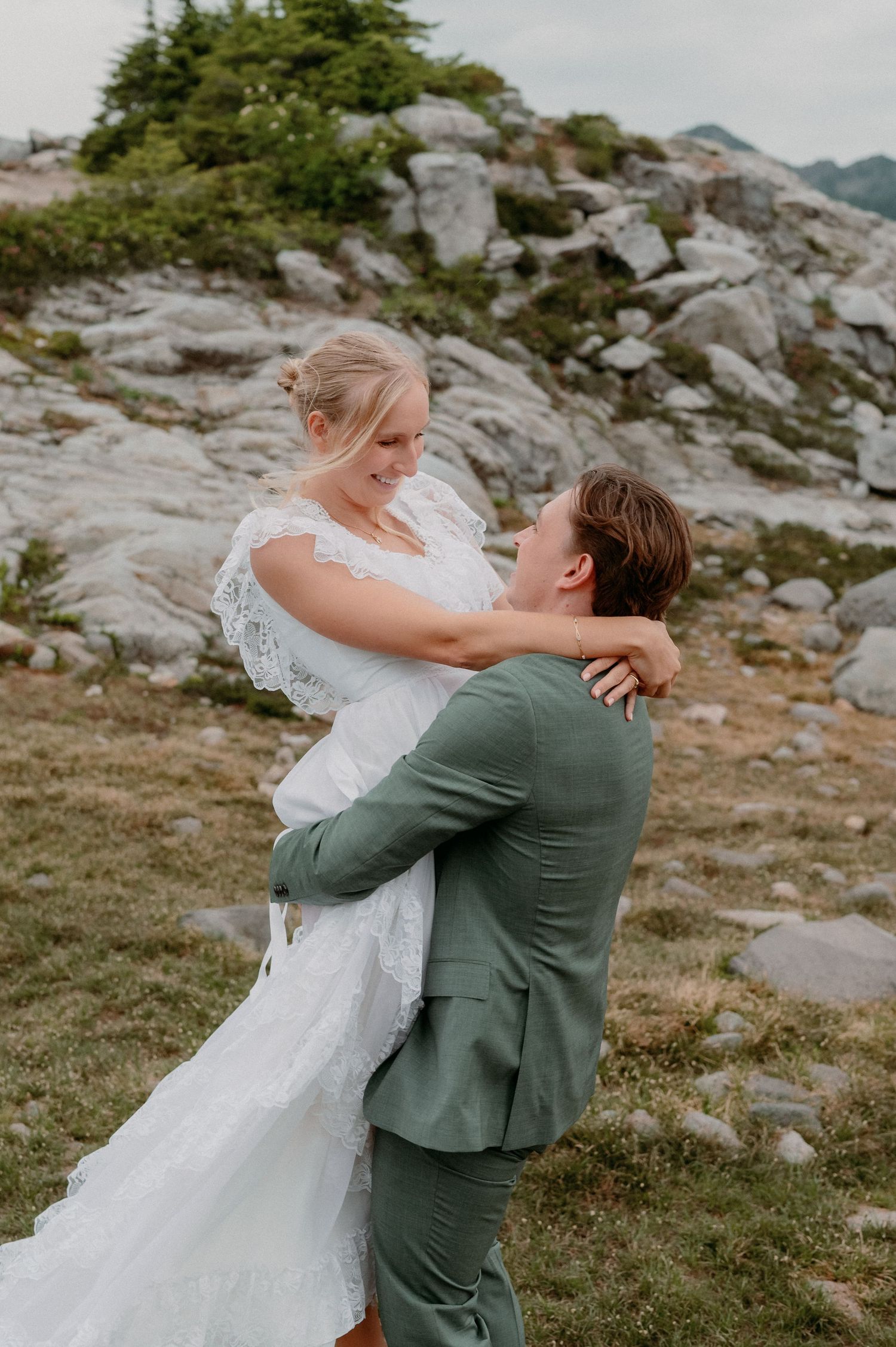 A romantic embrace between a couple in wedding attire on a rocky mountain peak during an outdoor ceremony.