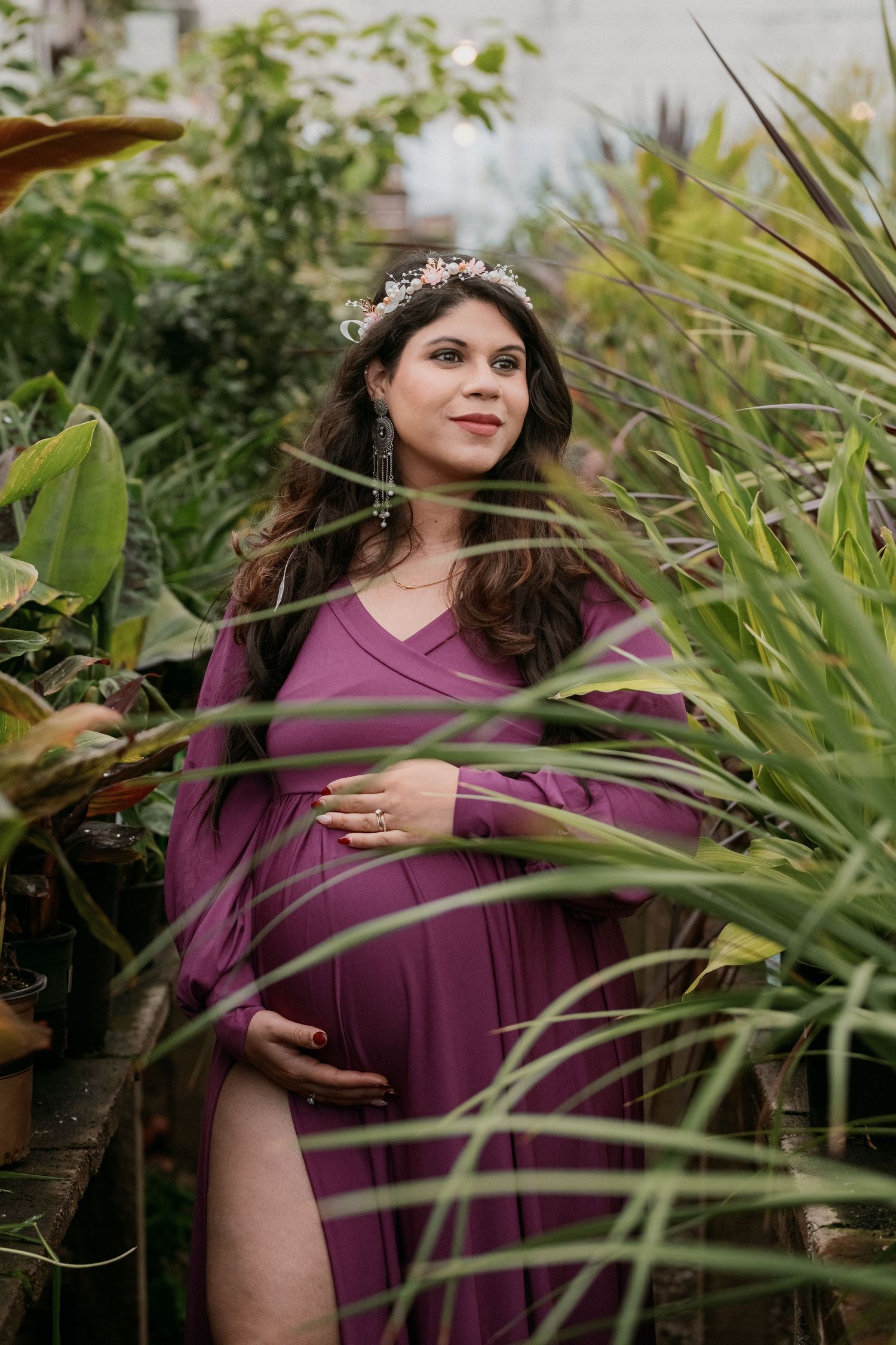 Expectant woman in purple dress stands among tropical plants in garden wearing delicate tiara.