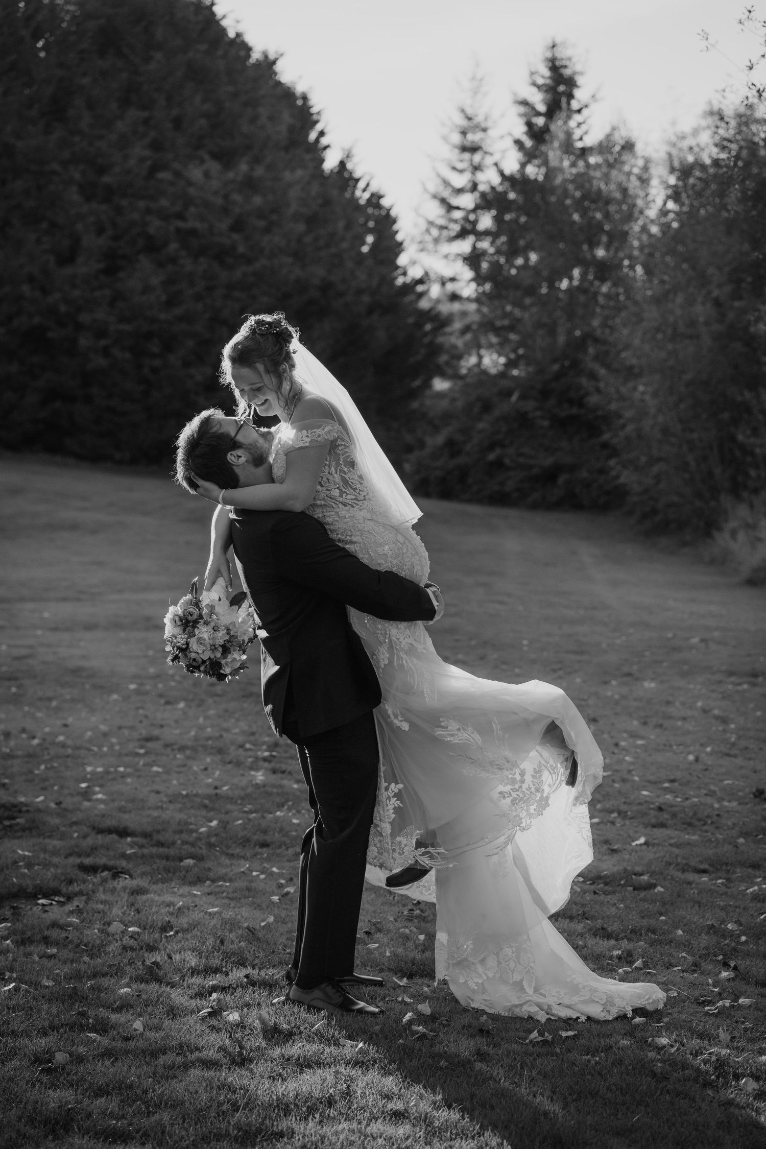 Black and white photo of romantic outdoor wedding moment with couple embracing in elegant formal attire in forest setting.