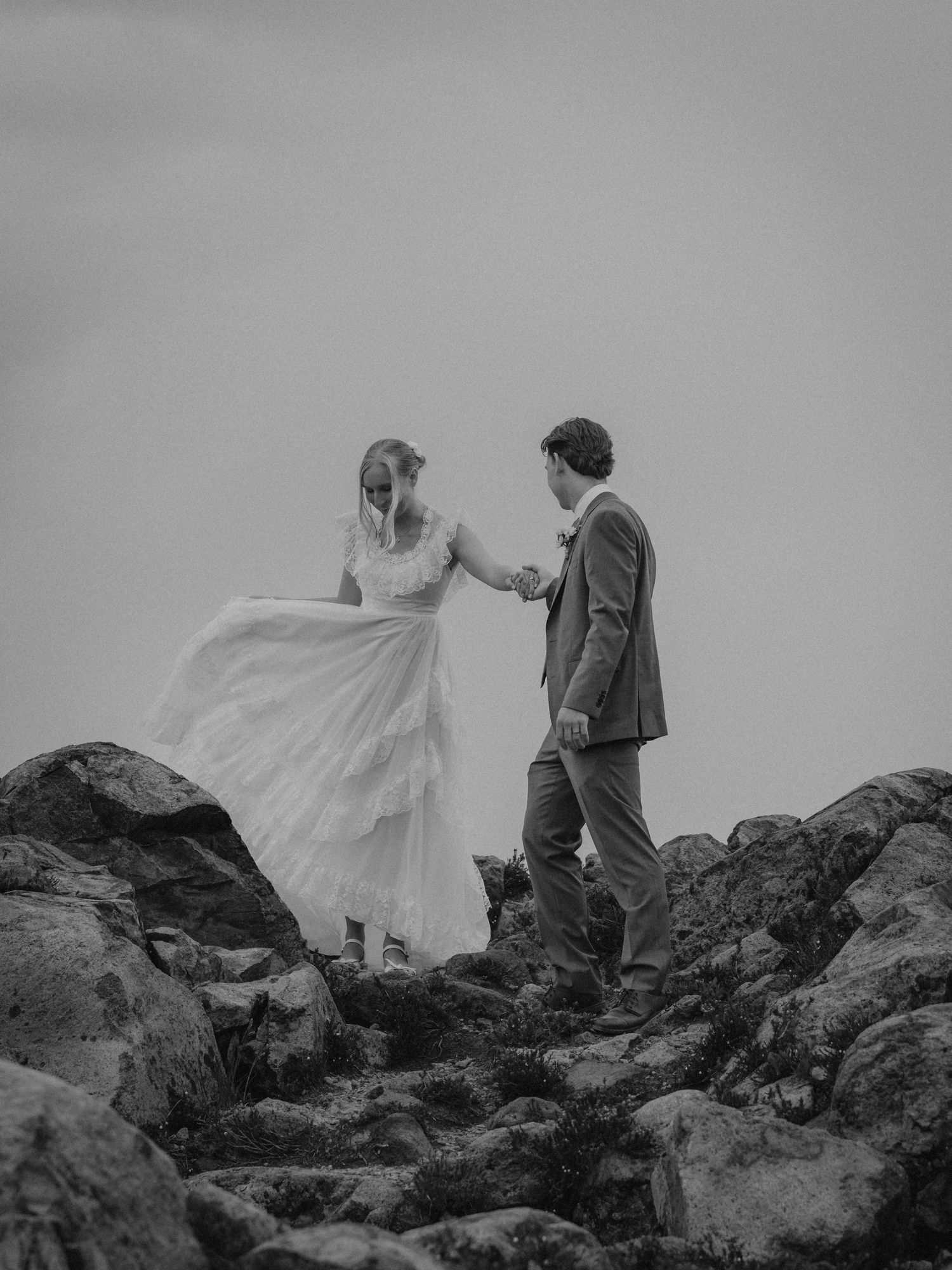 A bride and groom climb rocky terrain in black and white, with her flowing wedding dress trailing behind them.