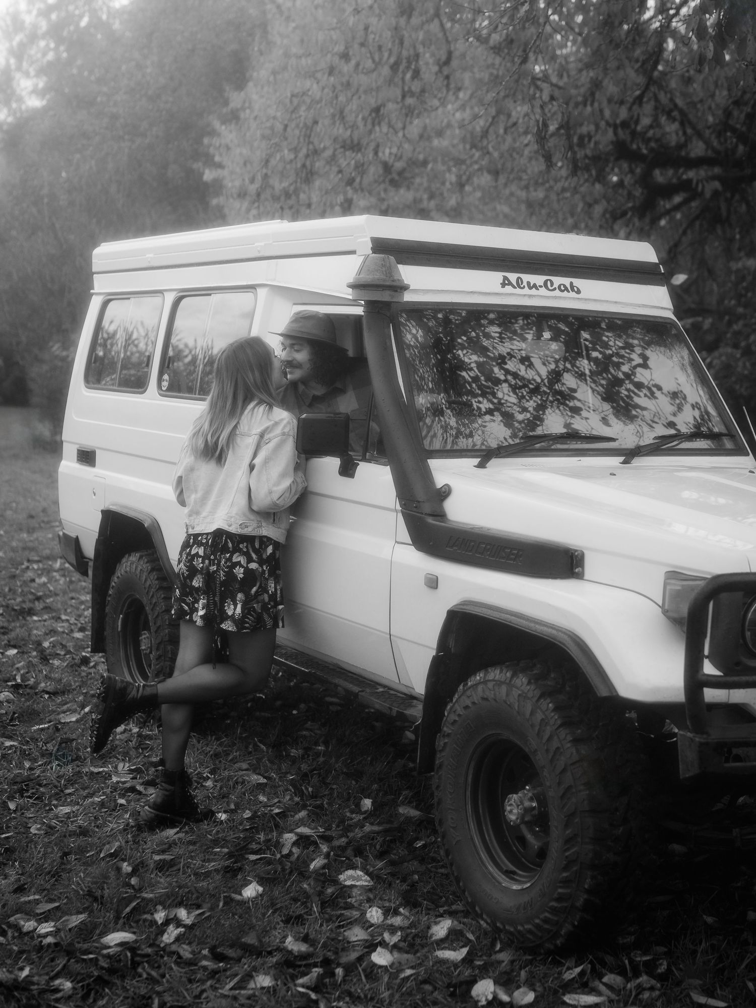 A black and white photo of someone leaning against a white Land Rover Defender off-road vehicle in a wooded area.