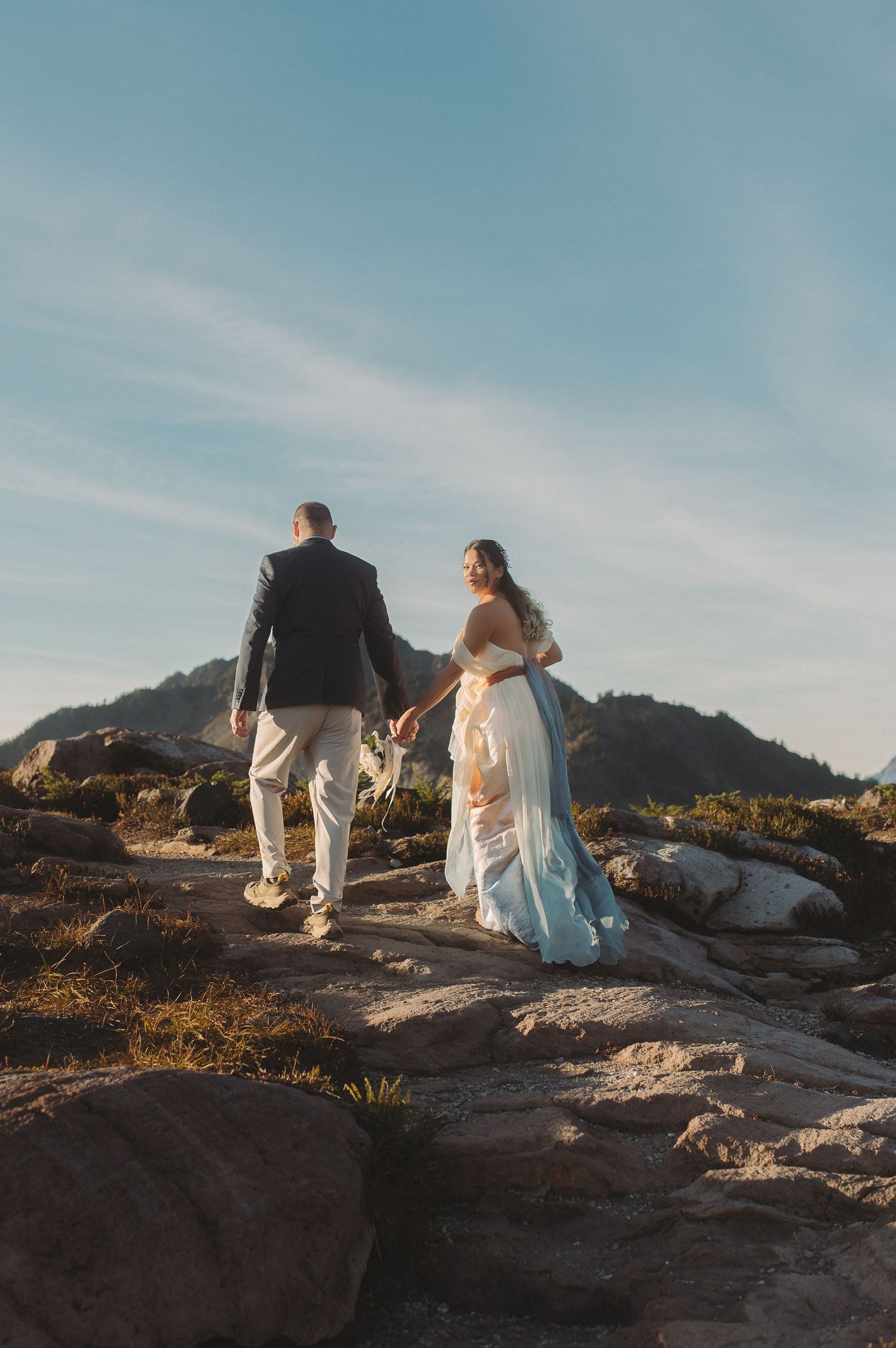 A couple walks along a rocky mountain path at sunset, silhouetted against a soft blue sky.