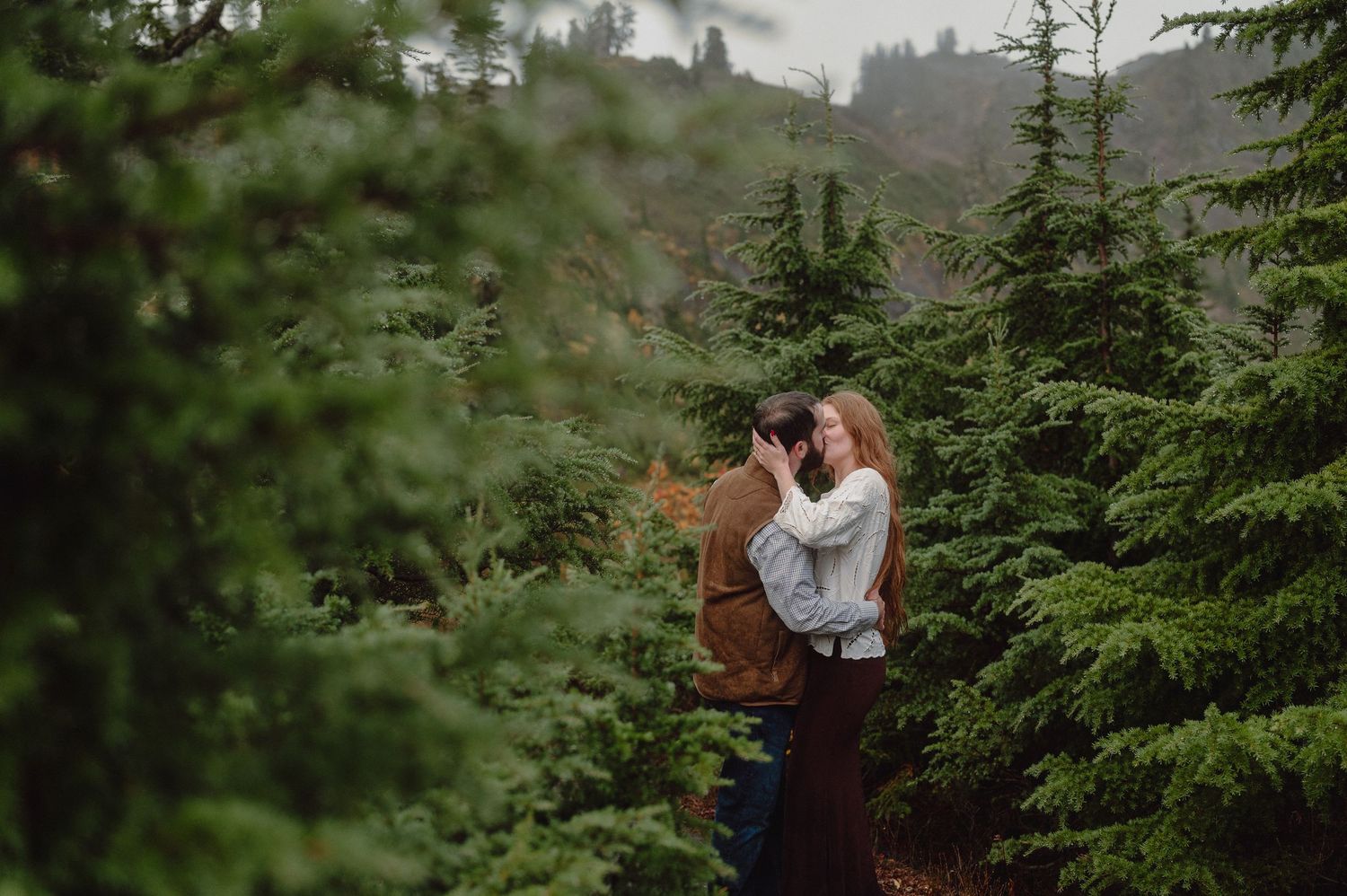 A couple stands among tall evergreen trees in a misty forest setting during an outdoor photography session.