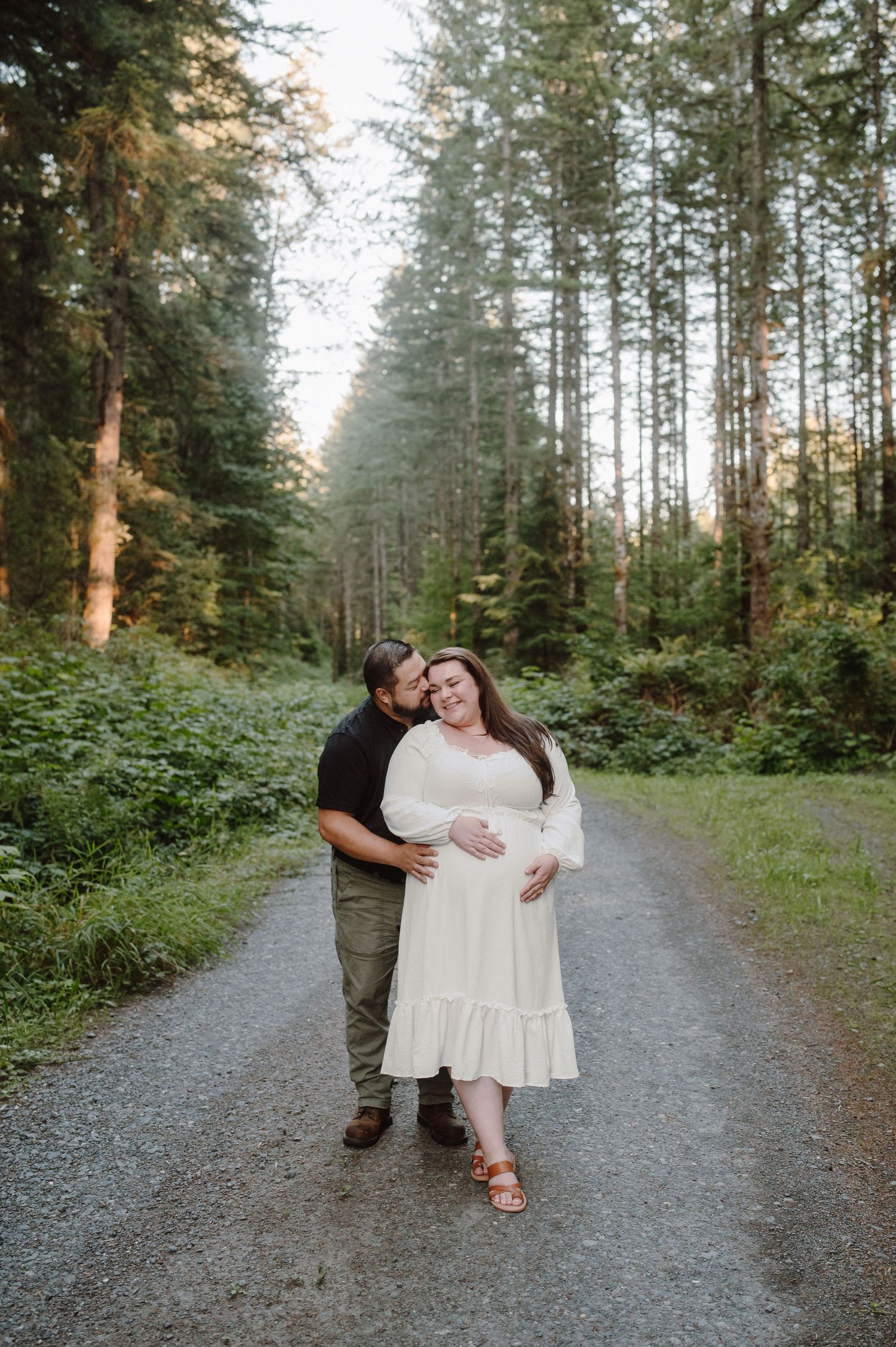 A couple embraces on a gravel path surrounded by tall evergreen trees in a misty Pacific Northwest forest.