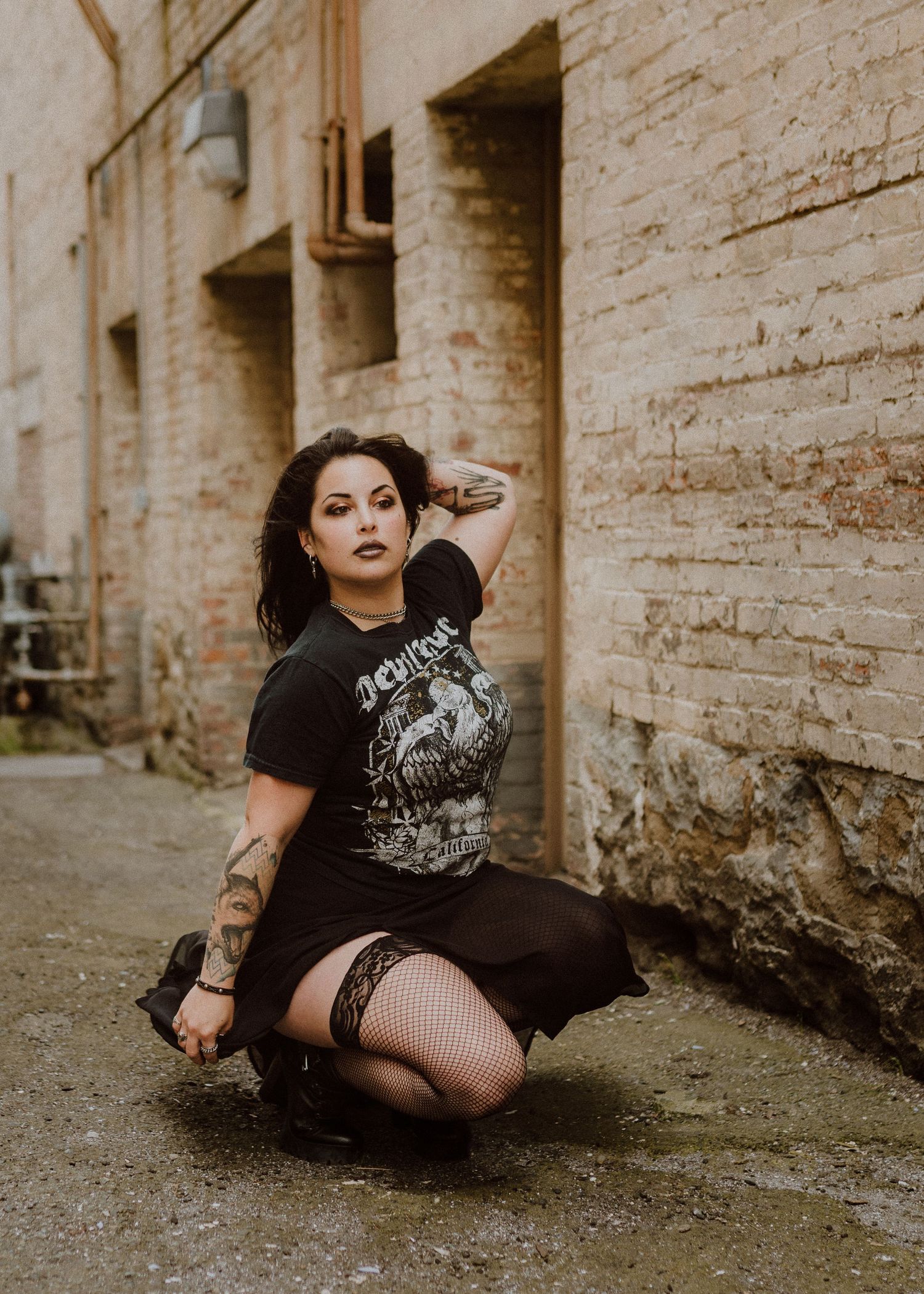 Alternative fashion model in black band shirt and fishnet stockings poses against brick wall in urban setting.