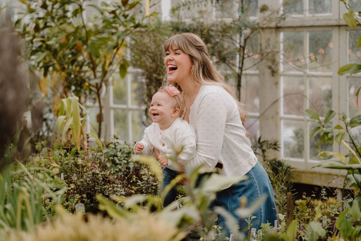 A joyful mother and baby laugh together among lush greenery inside a bright greenhouse.