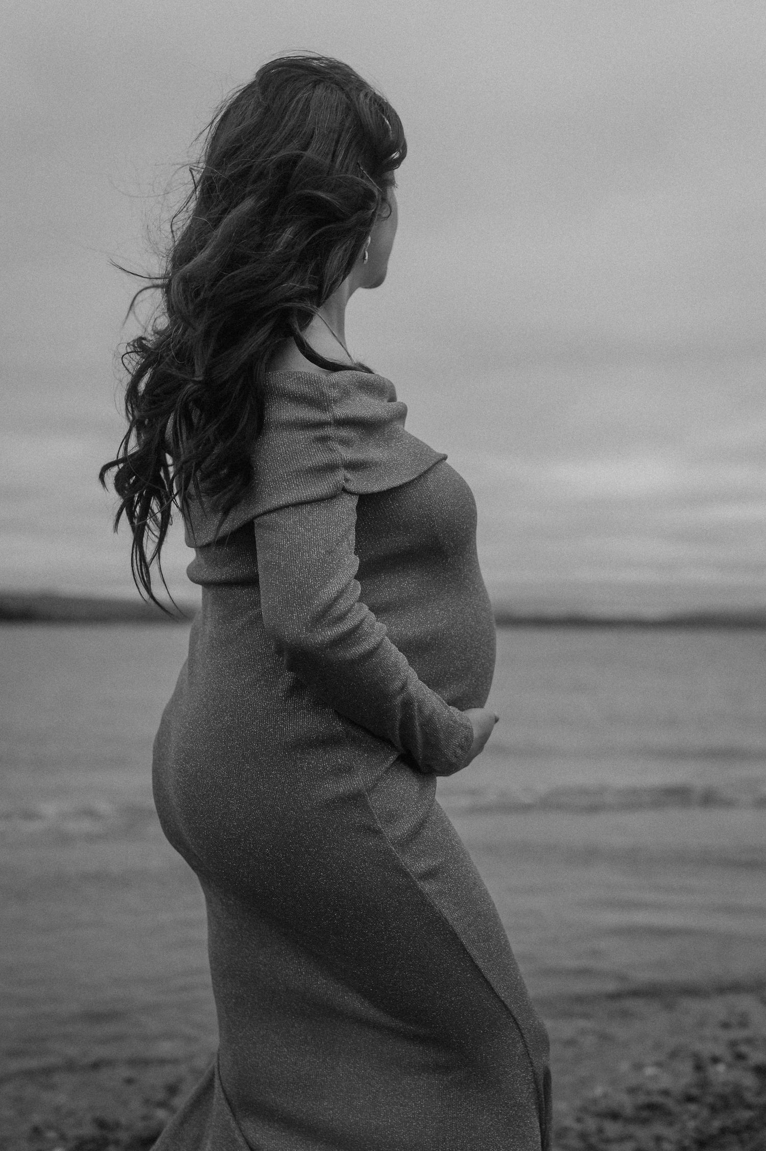 Black and white photo of pregnant woman in fitted dress standing by the ocean, cradling her baby bump.