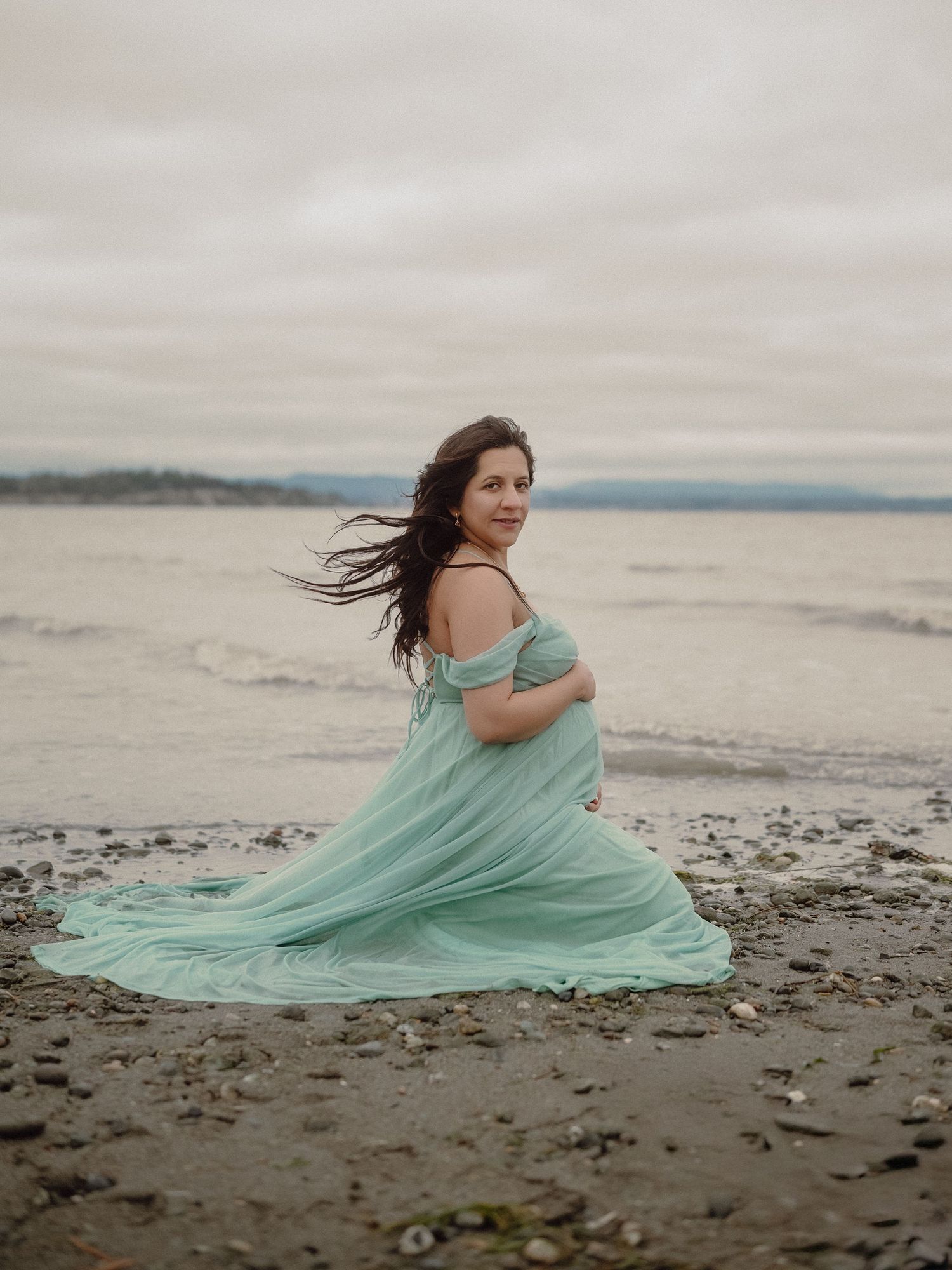 Pregnant woman in flowing mint green gown poses on rocky beach with ocean backdrop on overcast day.