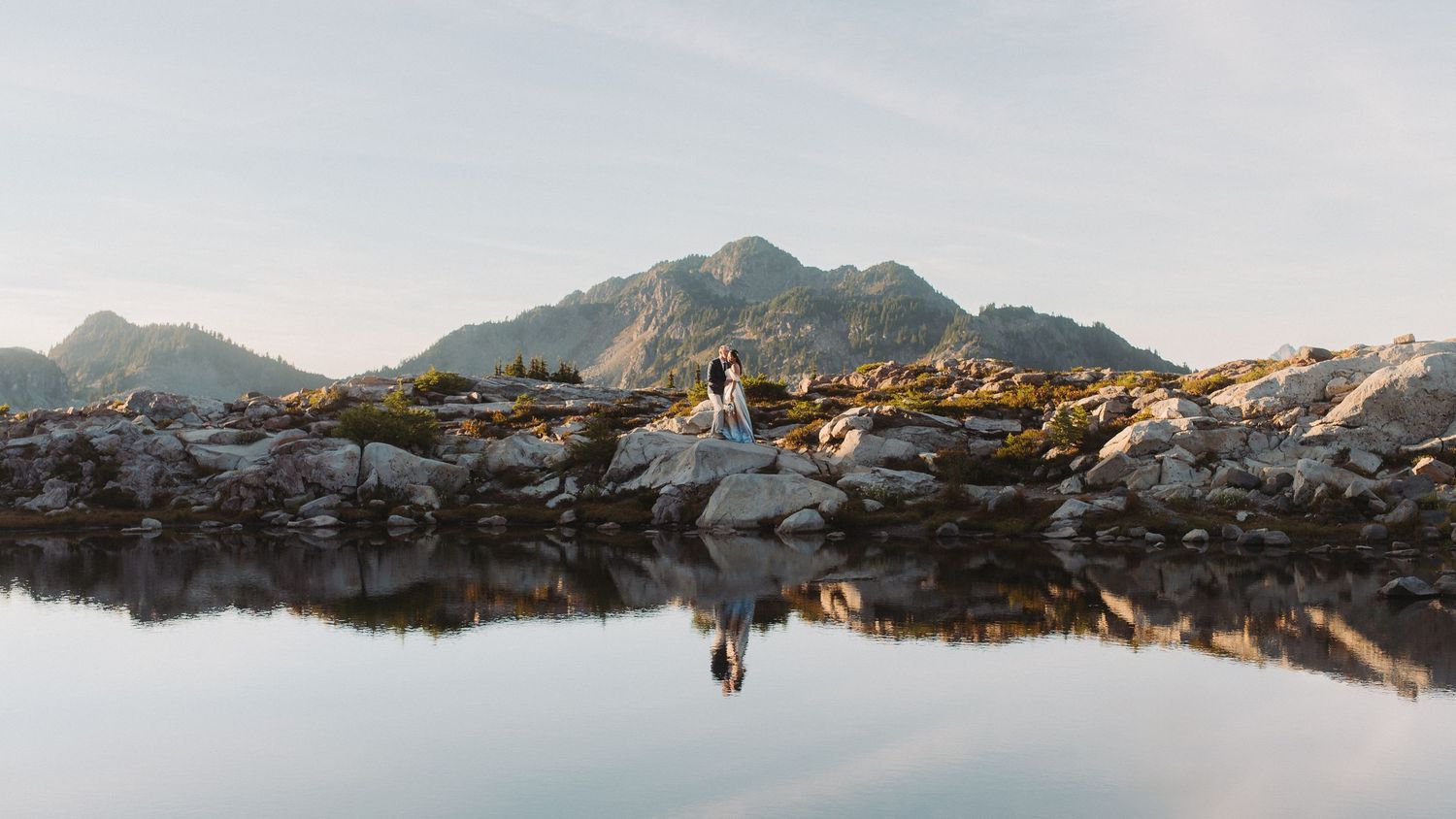 A panoramic view of a mountain lake reflecting snow-capped peaks and rugged terrain in golden sunlight.