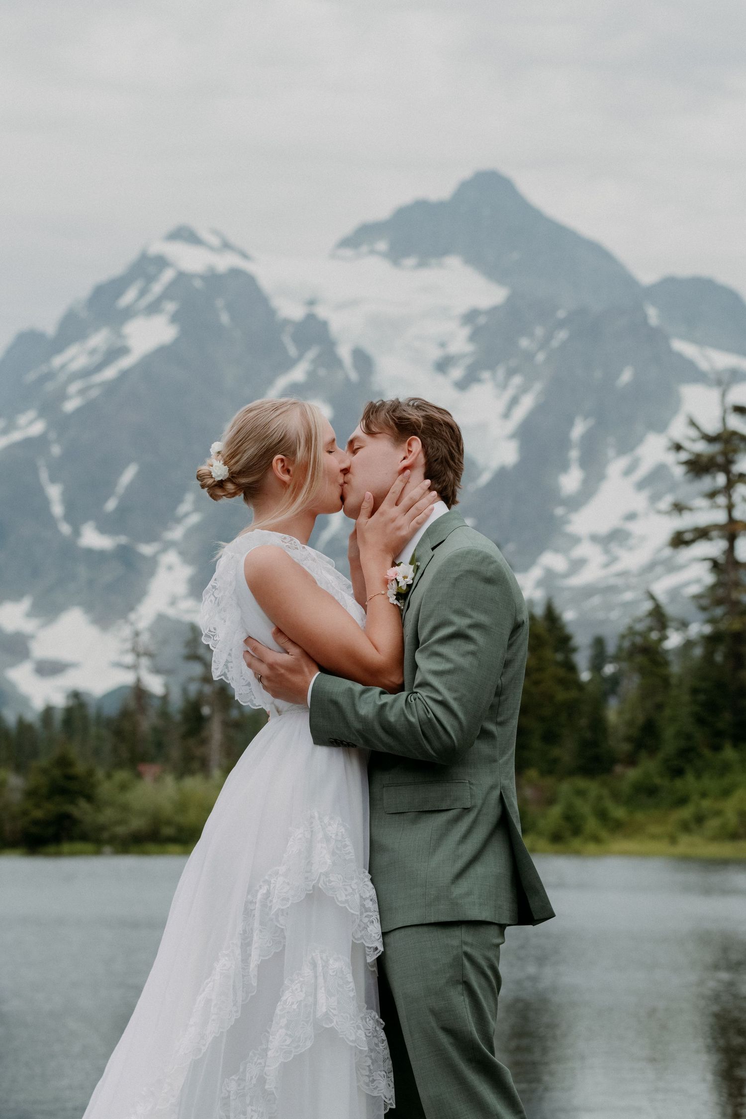 A romantic wedding couple shares a kiss against a stunning backdrop of snow-capped mountains and a serene lake.