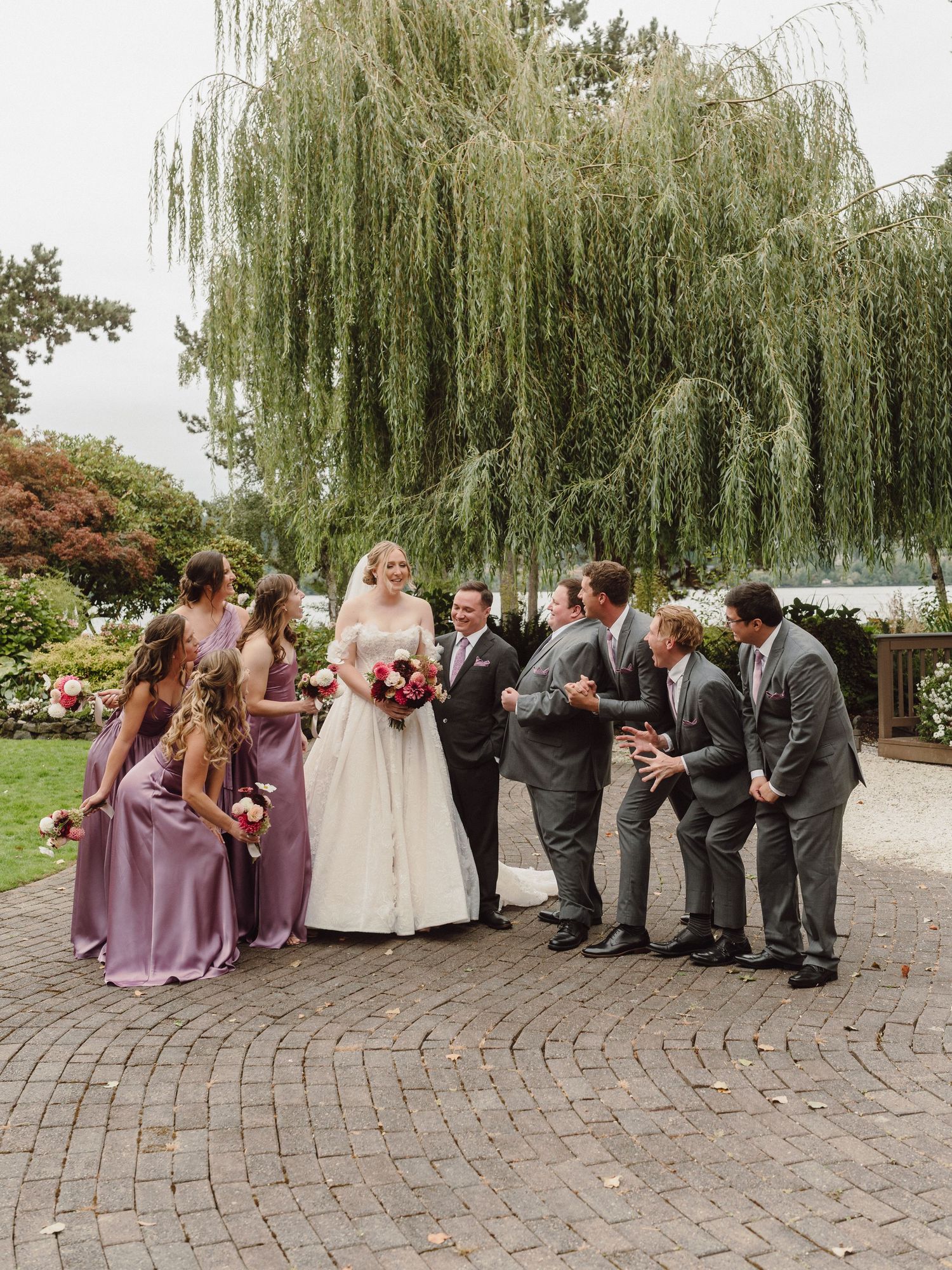 Wedding party poses on brick patio with elegant willow tree and garden backdrop during outdoor celebration.