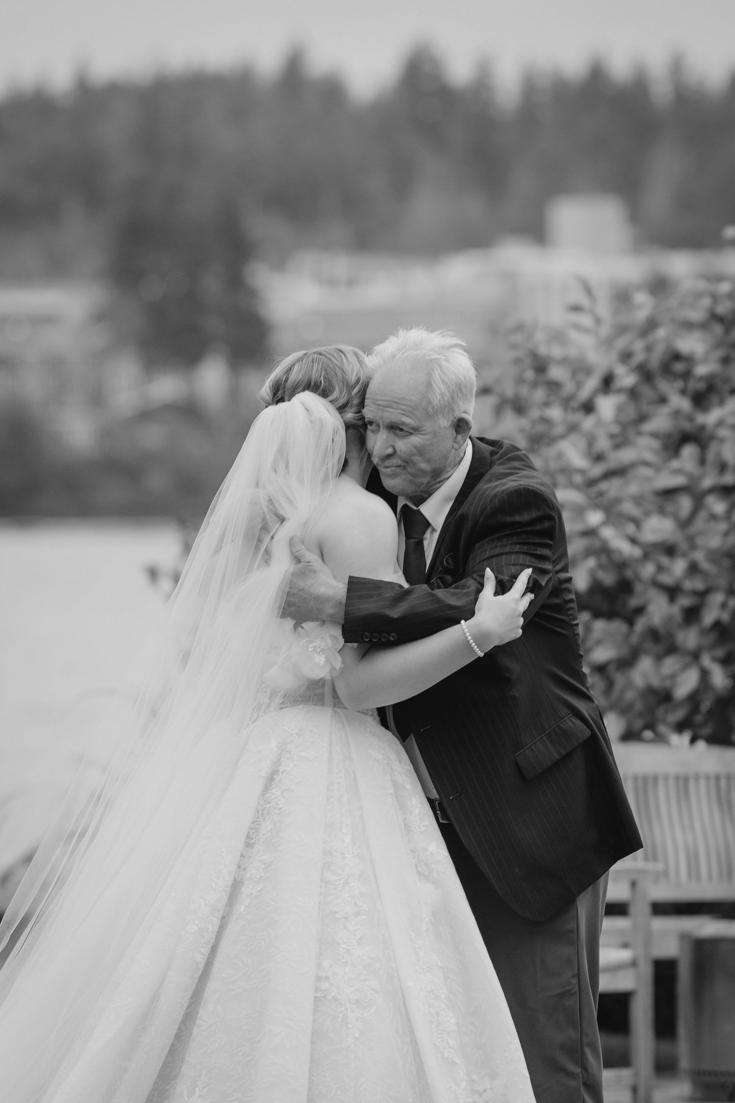 Black and white photo of a bride and older man sharing an emotional hug at an outdoor wedding ceremony.