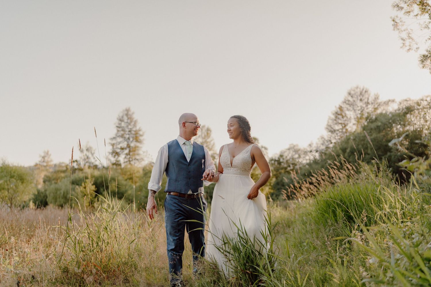 Newlyweds walk hand in hand through a golden meadow during sunset.