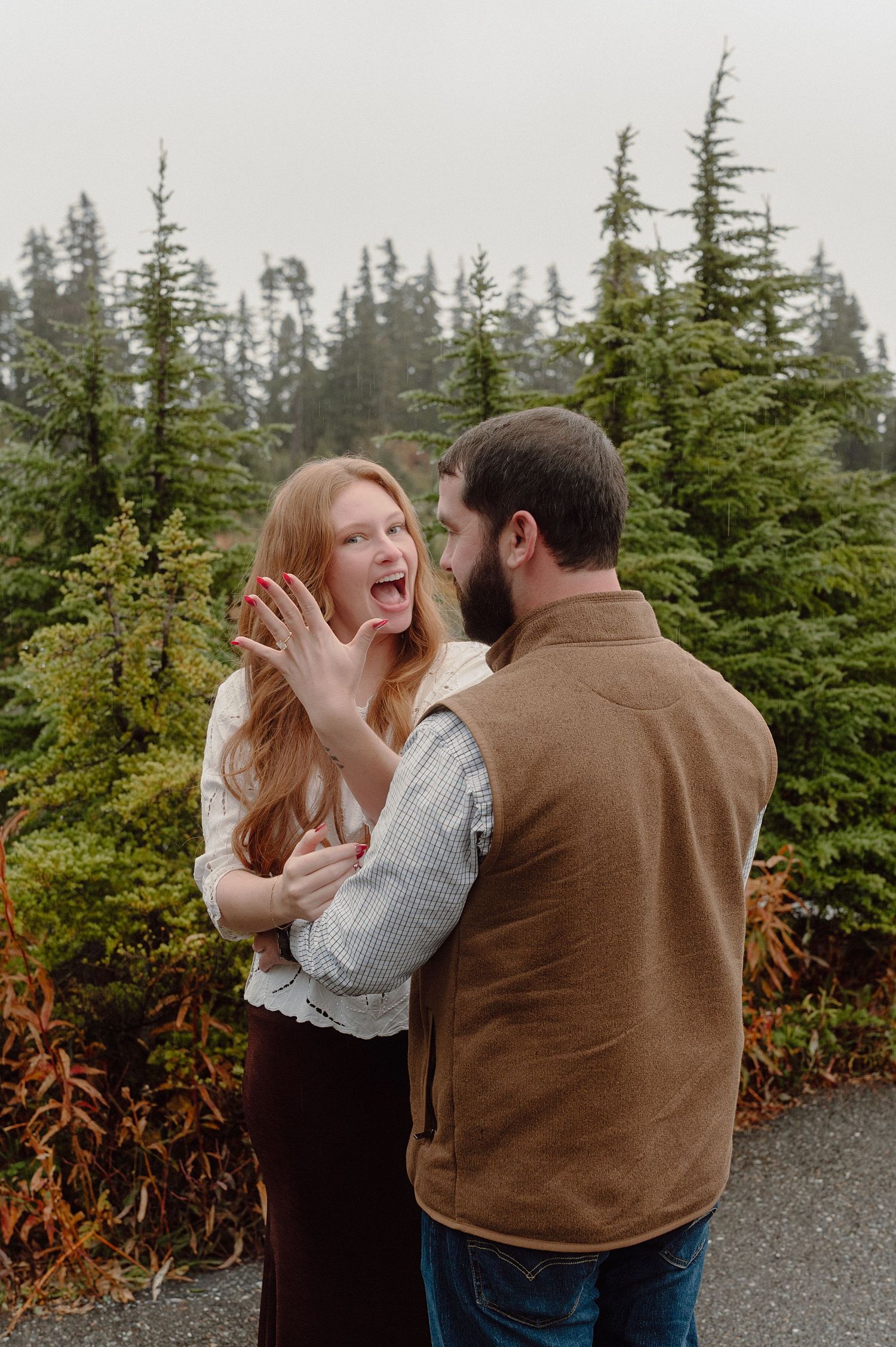 A couple shares a tender moment together among evergreen trees on a misty mountain path.