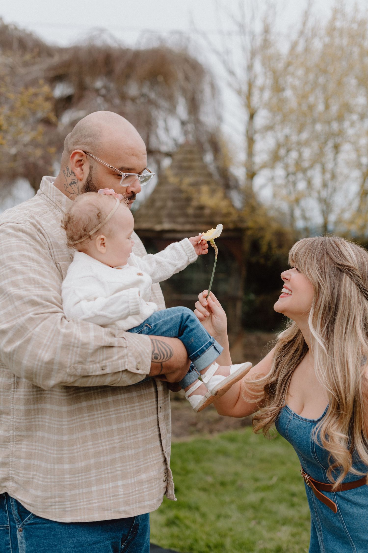A bald father holds a baby offering a flower to a smiling blonde woman outdoors in a garden setting.