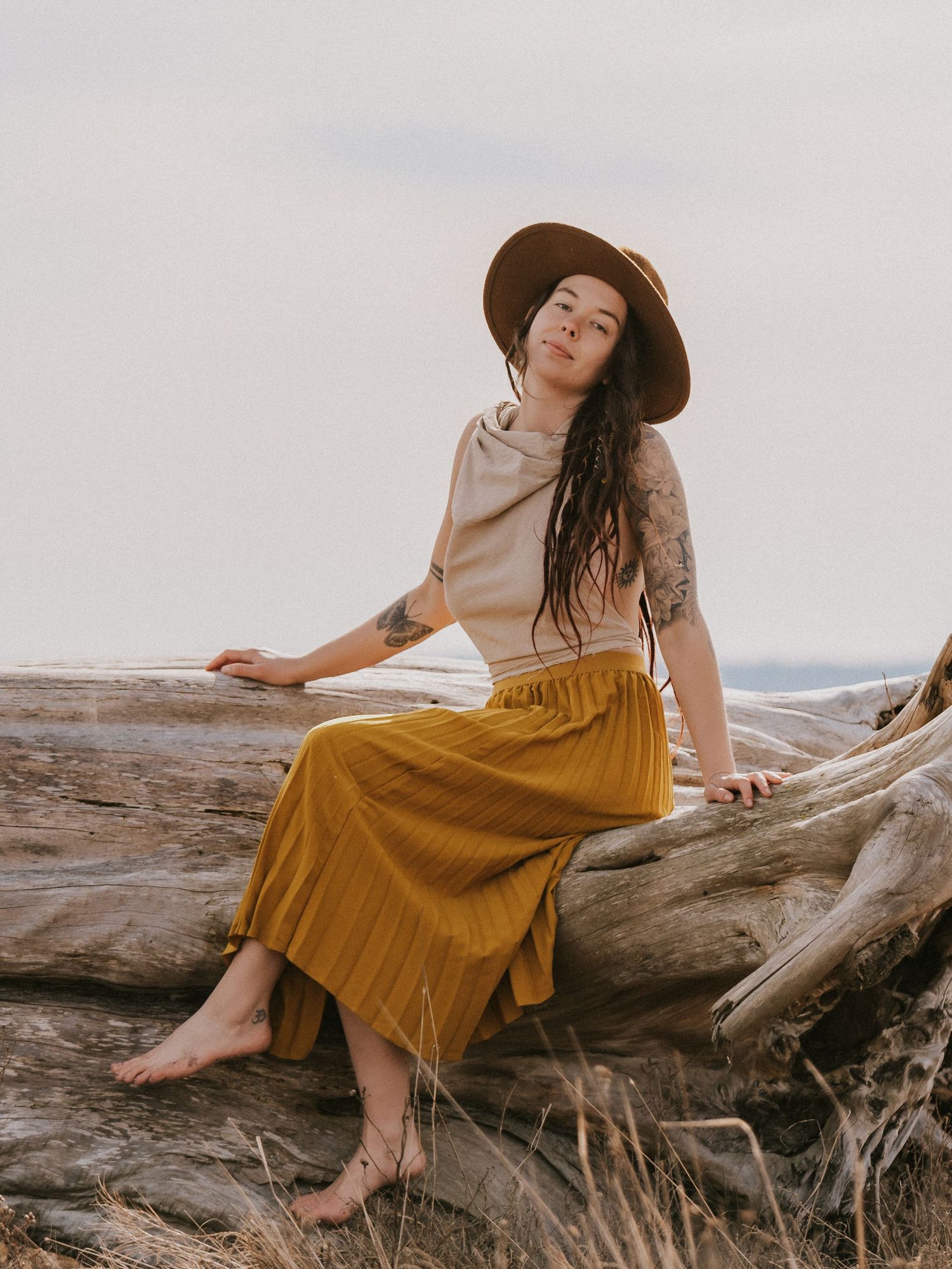 Tattooed woman in mustard skirt and brown hat sits barefoot on driftwood in a coastal outdoor setting.