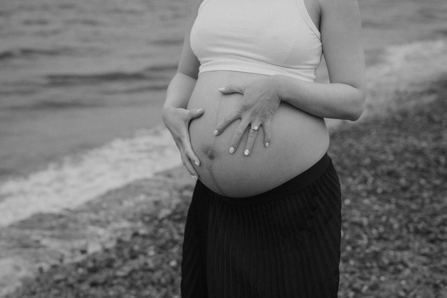 Close-up black and white shot of hands cradling a pregnant belly by the ocean.
