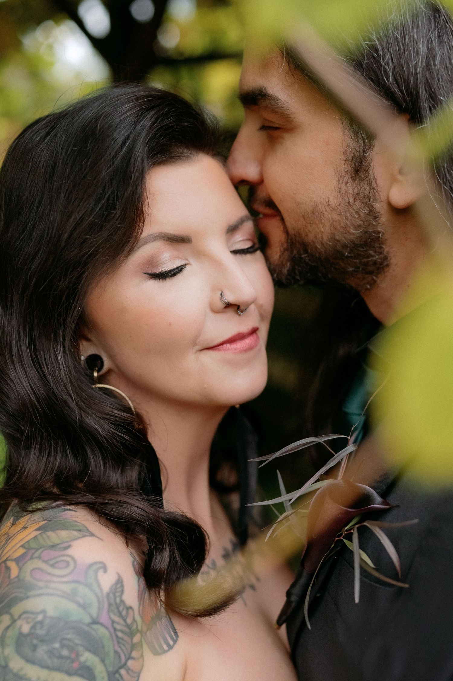 Intimate romantic portrait of a couple among green foliage, with visible tattoos and gentle expressions.