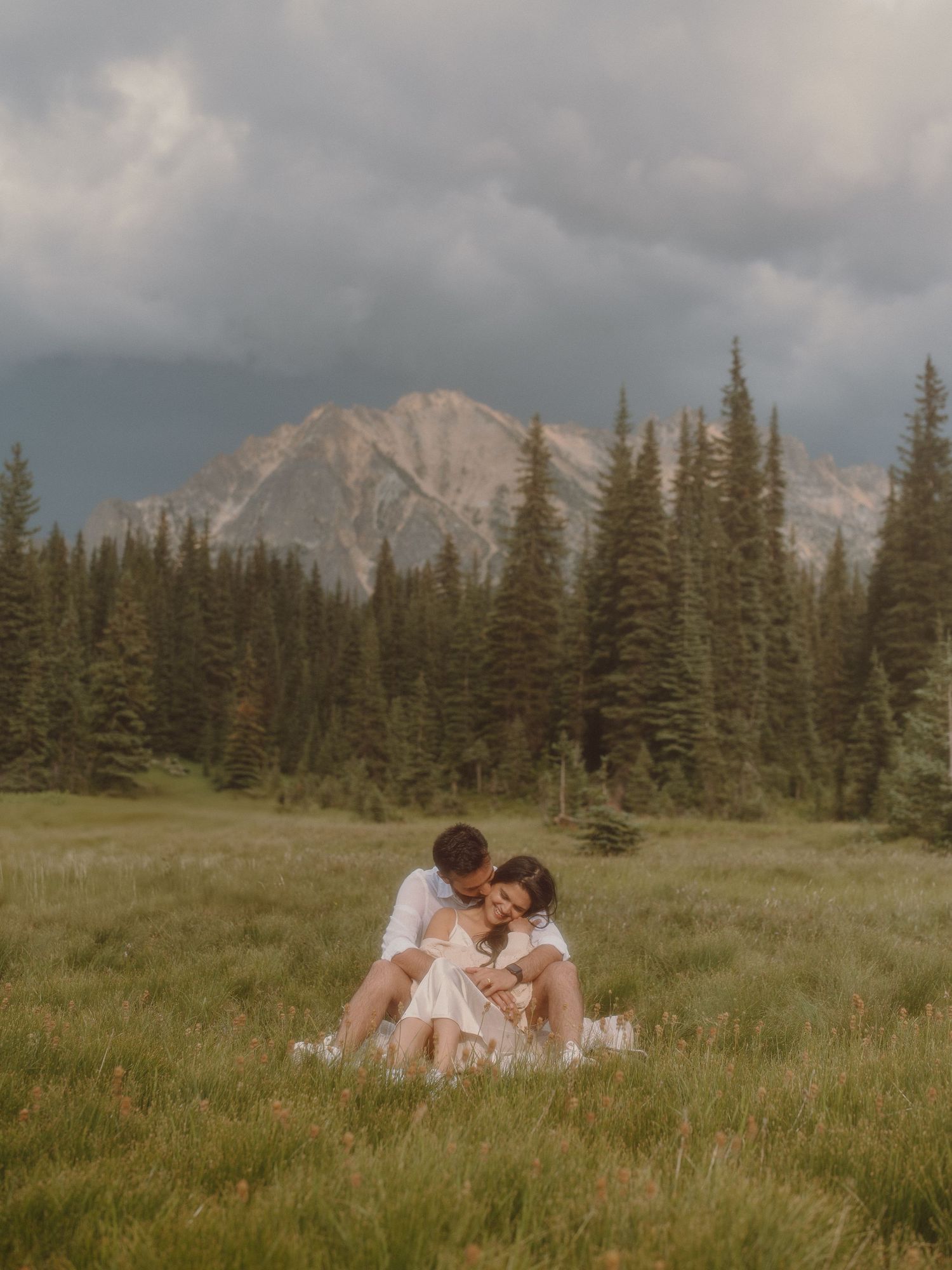 A couple sharing an intimate moment in a mountain meadow with dramatic peaks in the background.