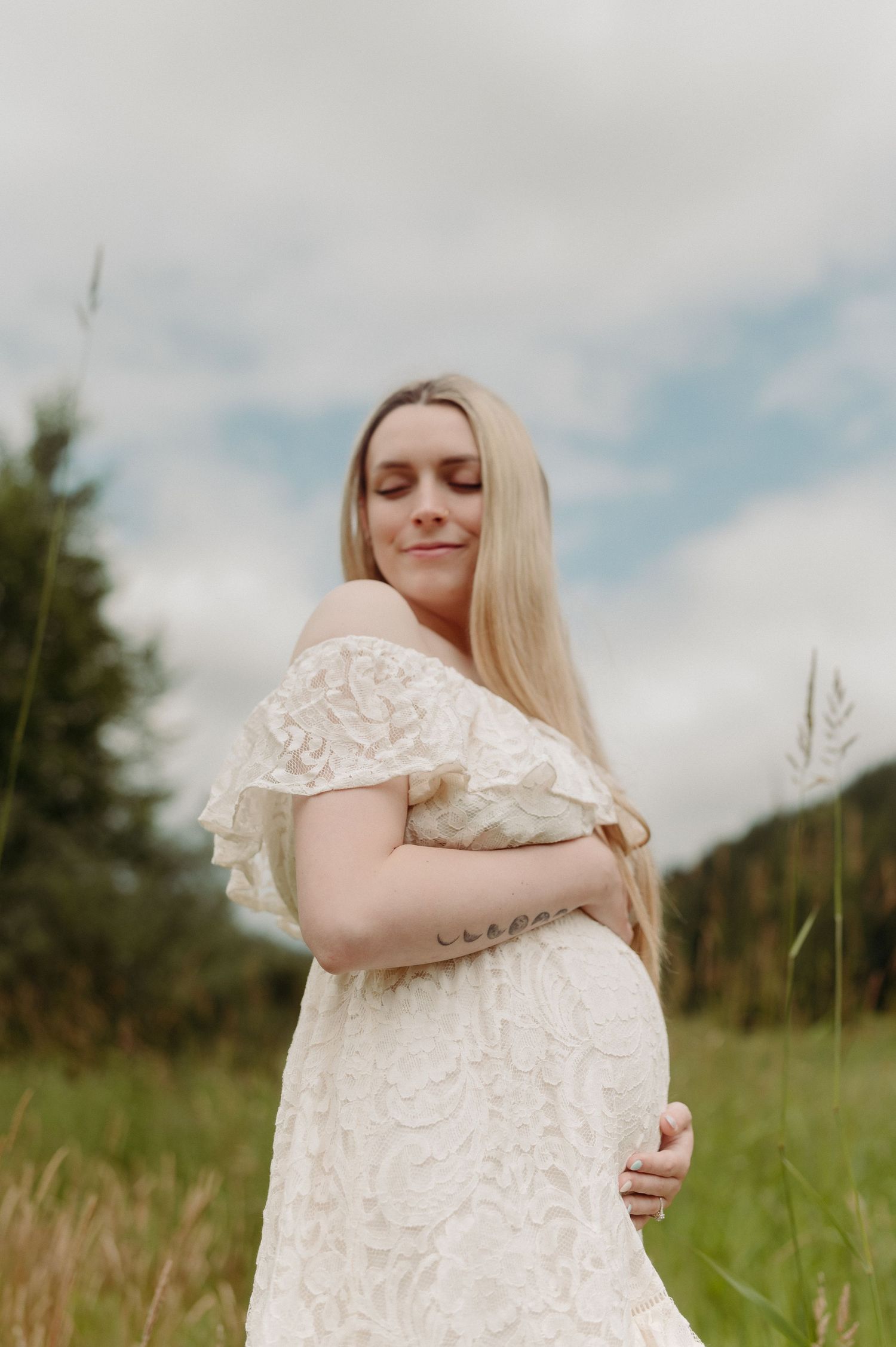 Pregnant woman in white lace off-shoulder dress standing in a field, cradling her baby bump with eyes closed.