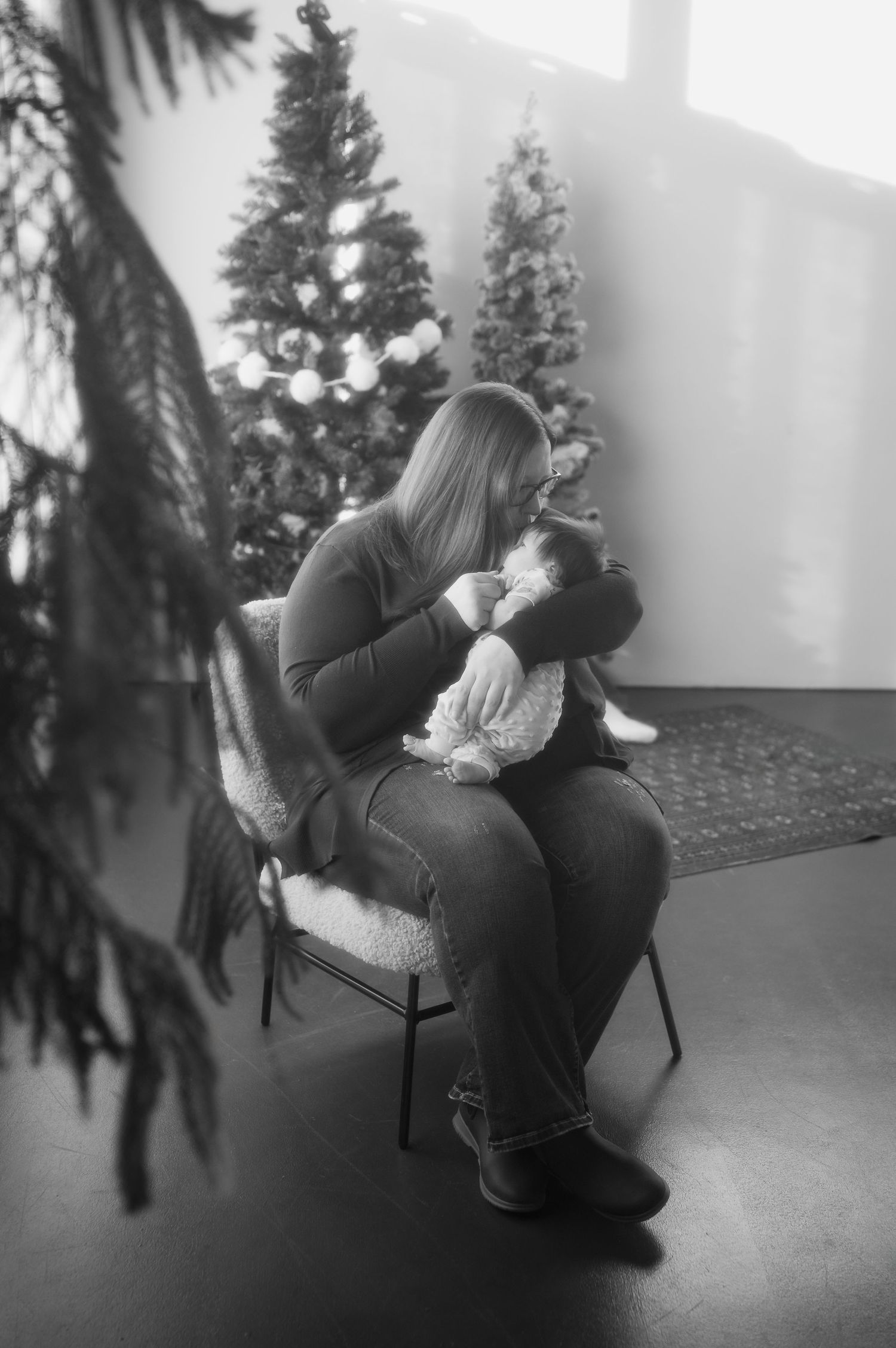Black and white photograph showing an intimate moment between parent and child in a chair beside a Christmas tree.