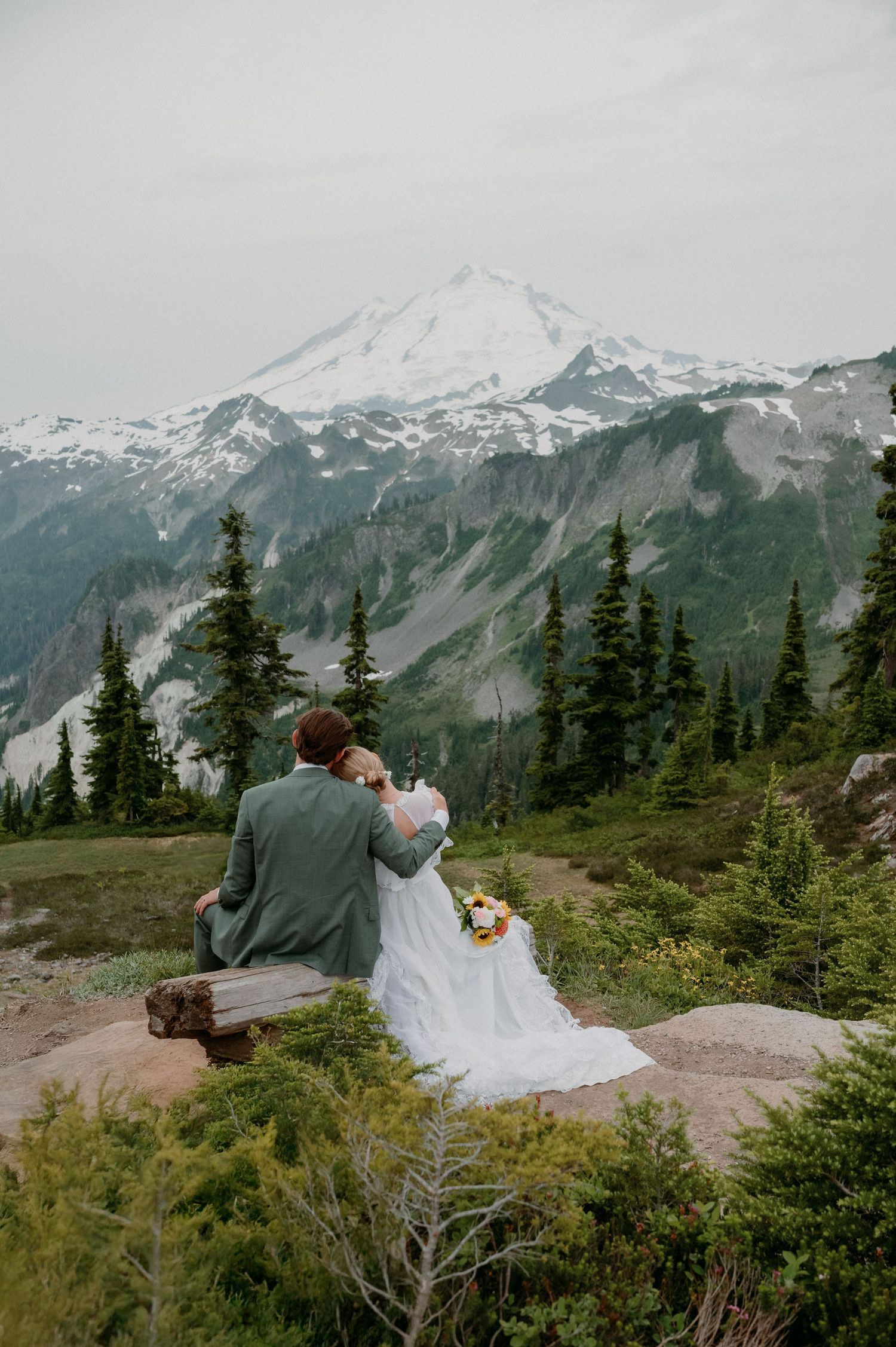 A bride and groom sit on a rocky overlook with a snow-capped mountain and evergreen forest backdrop during their elopement.