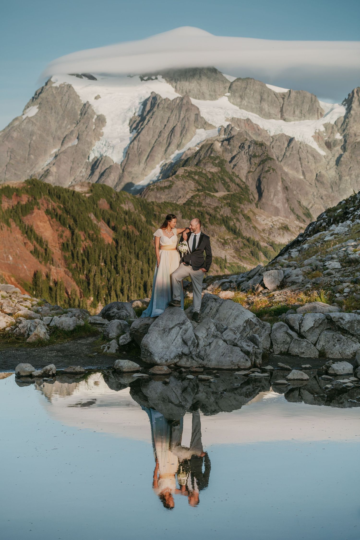 Couple standing on rocky mountain ledge overlooking snow-capped peaks in dramatic mountain landscape at sunset.