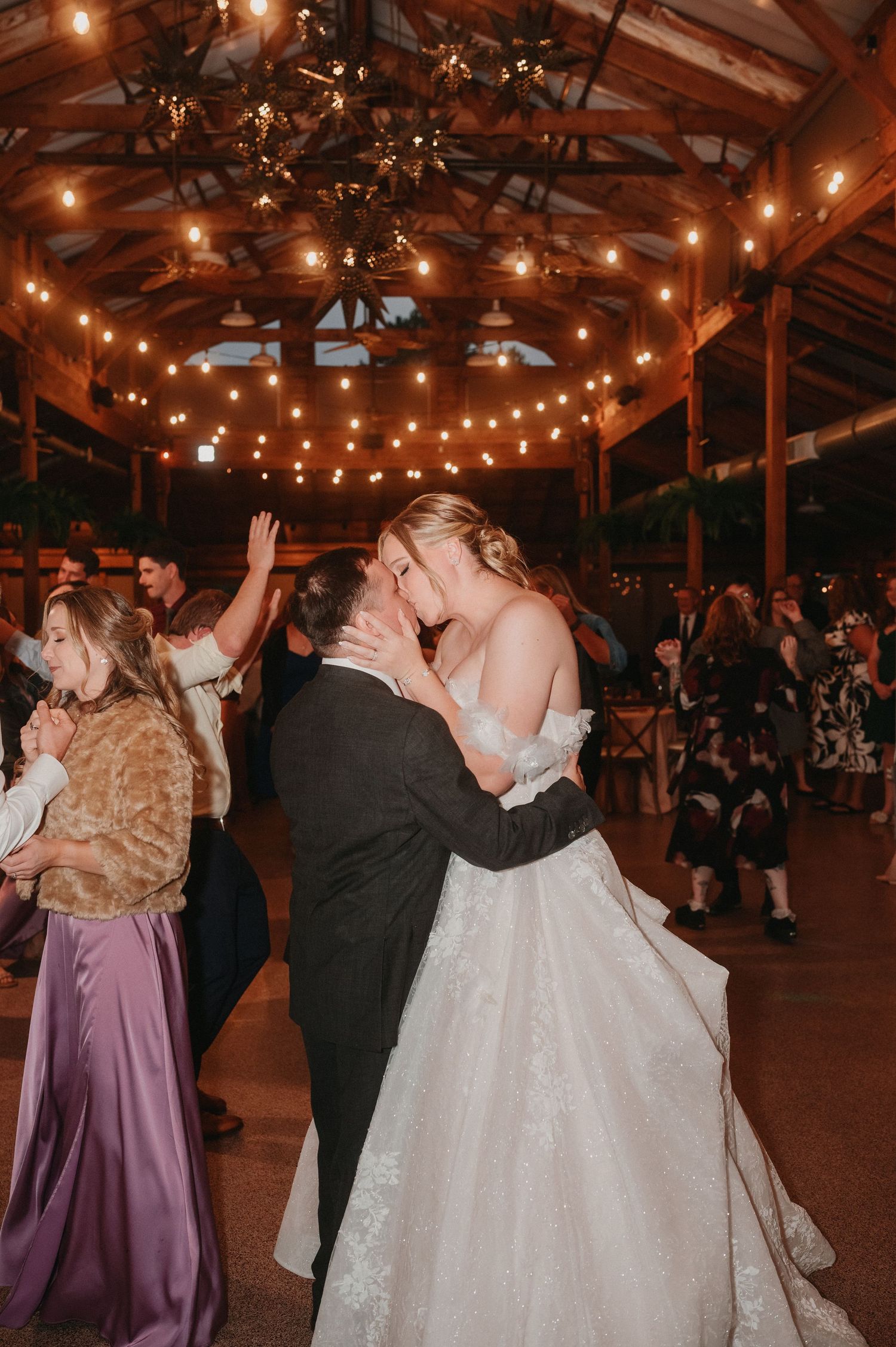 Wedding couple and guests dance together during romantic reception with glowing string lights overhead in rustic venue.