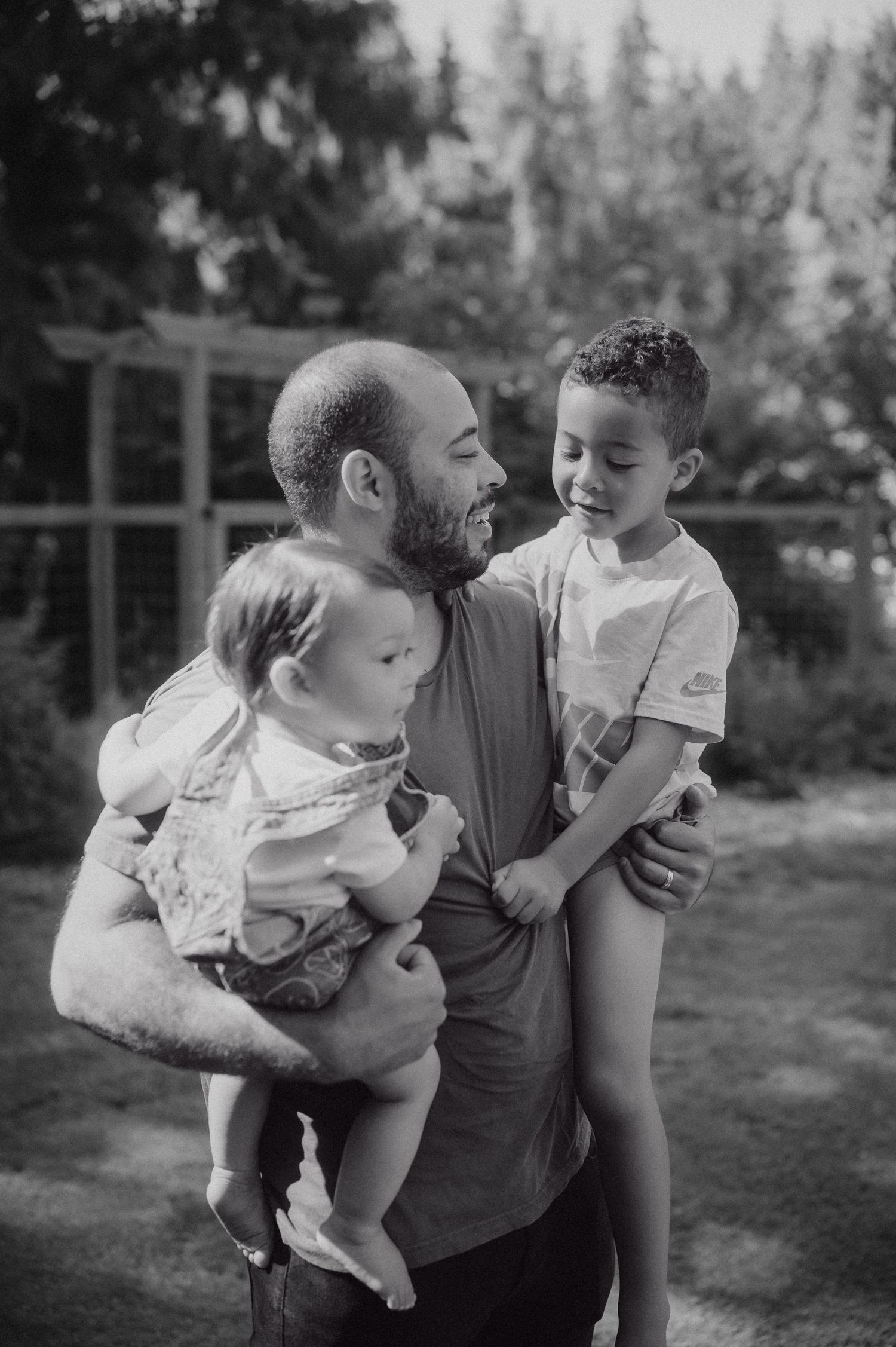 Black and white photo of a bearded father holding two young children outdoors, smiling warmly in a garden setting.
