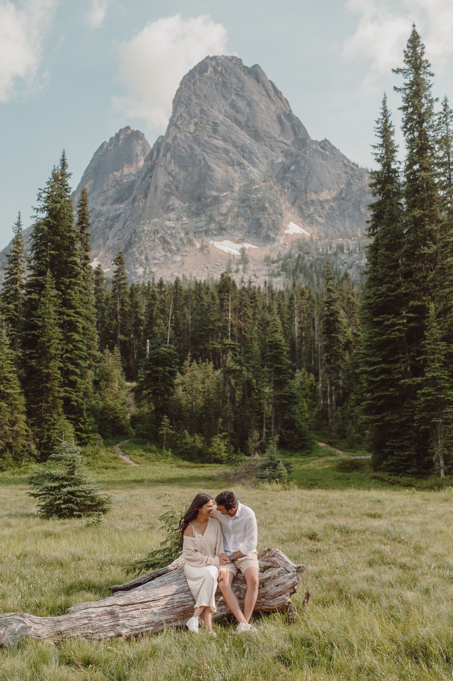 A couple sitting together on a fallen log with dramatic mountain peaks and pine forest in the background.
