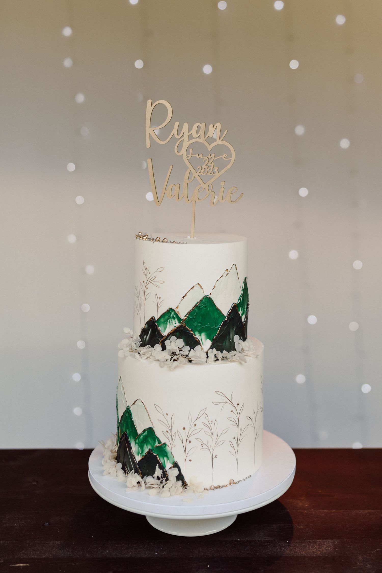 A two-tiered wedding cake decorated with green mountain designs and a personalized gold topper.