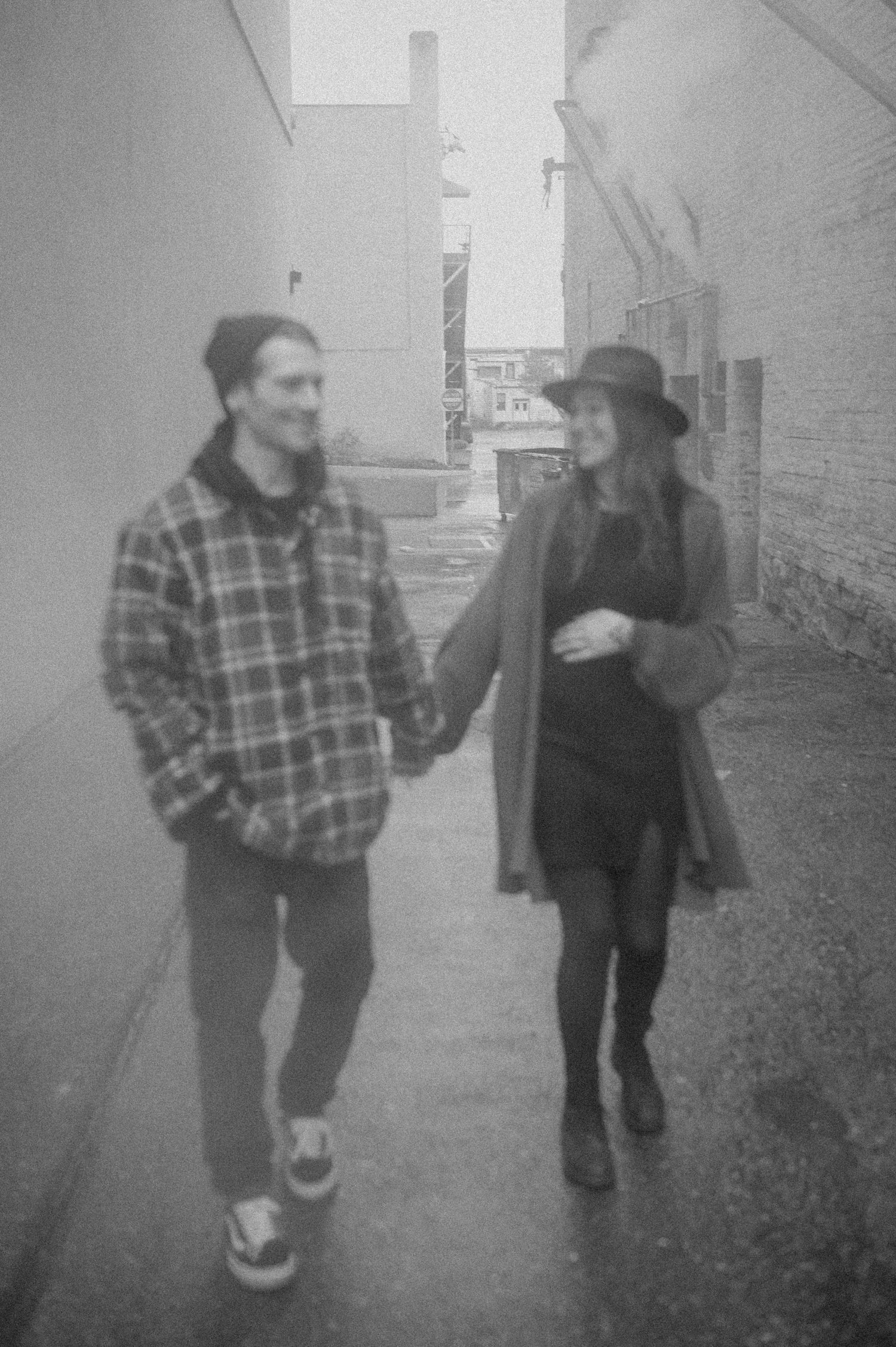 A couple walking together in casual attire down a misty alley in black and white.