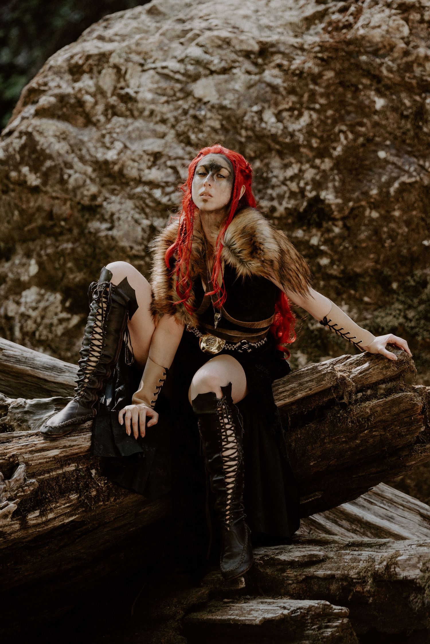 A woman in Viking warrior cosplay with red hair, fur collar, black outfit, and lace-up boots sits on a log near rocks.