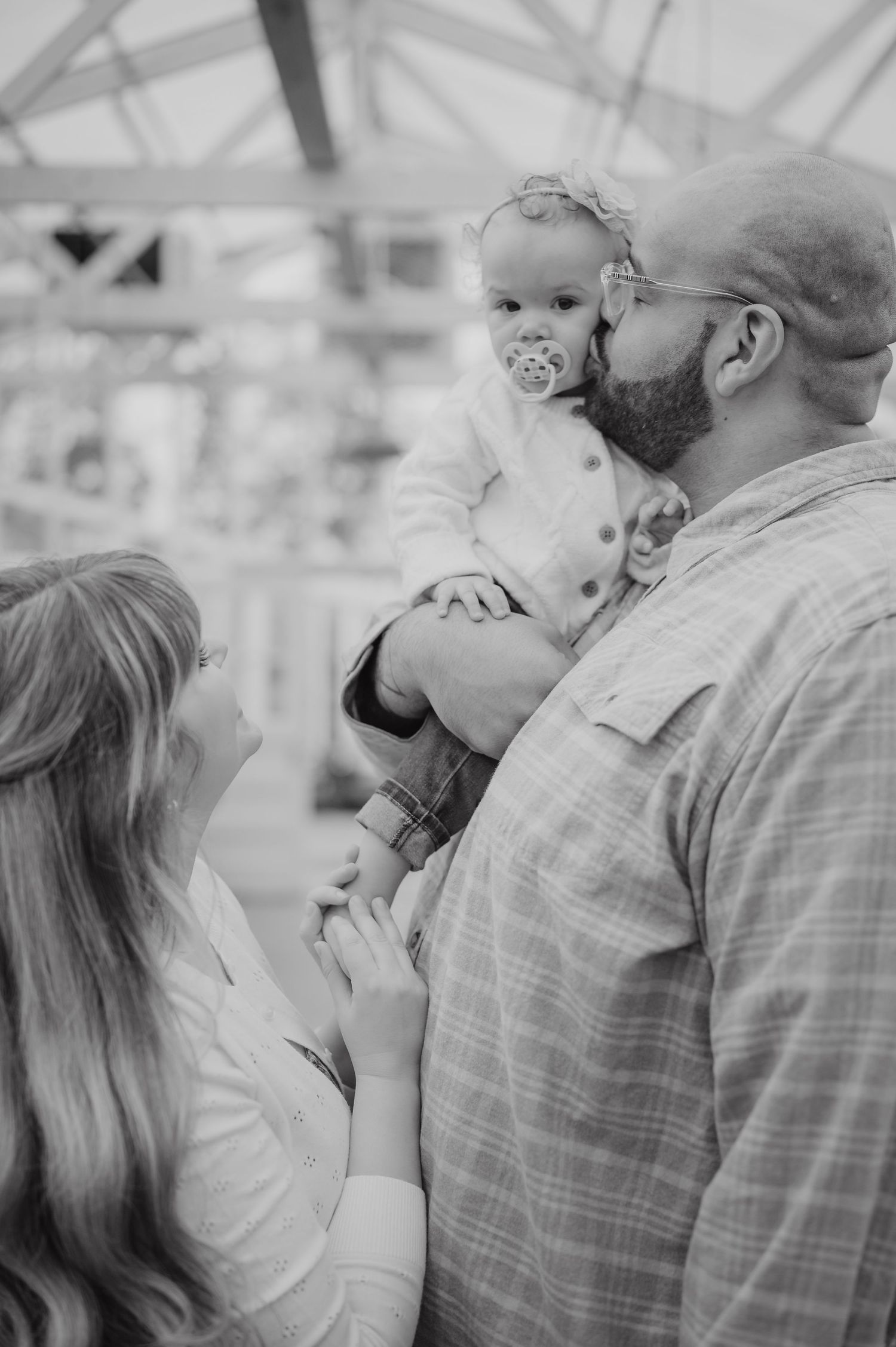 Black and white photo of a bald father kissing his baby while mother looks up lovingly in a greenhouse setting.