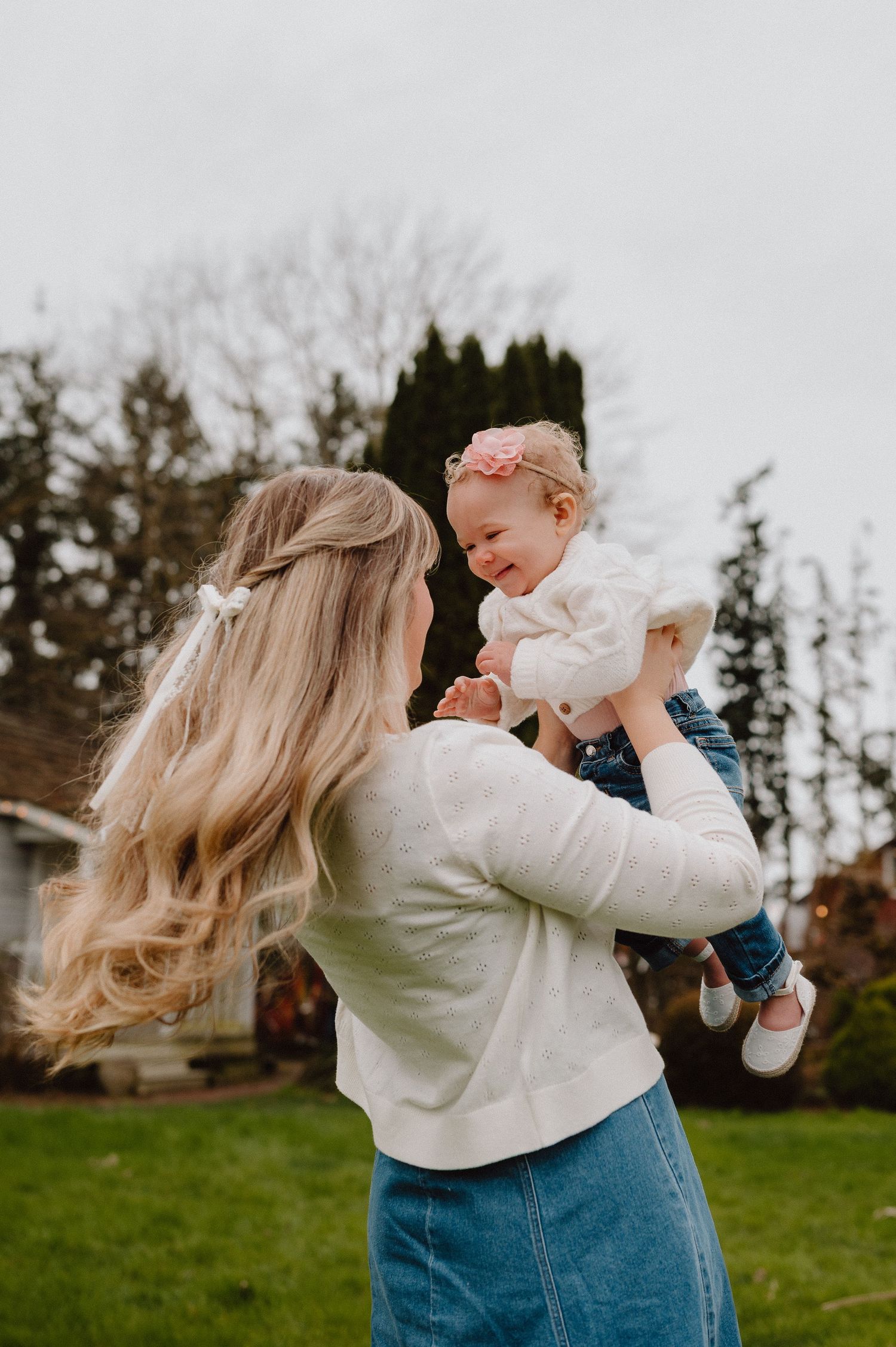 A blonde mother lifts her smiling baby girl wearing a pink headband outdoors on a green lawn.