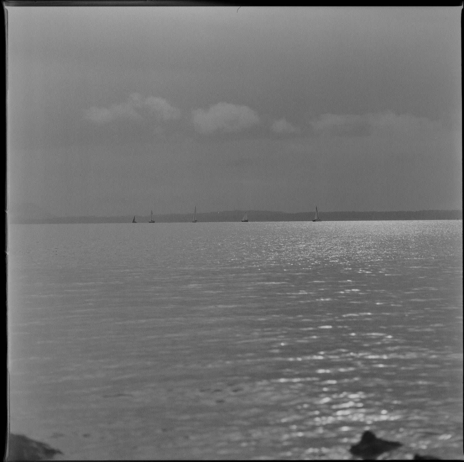 Grayscale seascape with dark silhouetted rocks visible in the foreground against a textured ocean and cloudy sky.