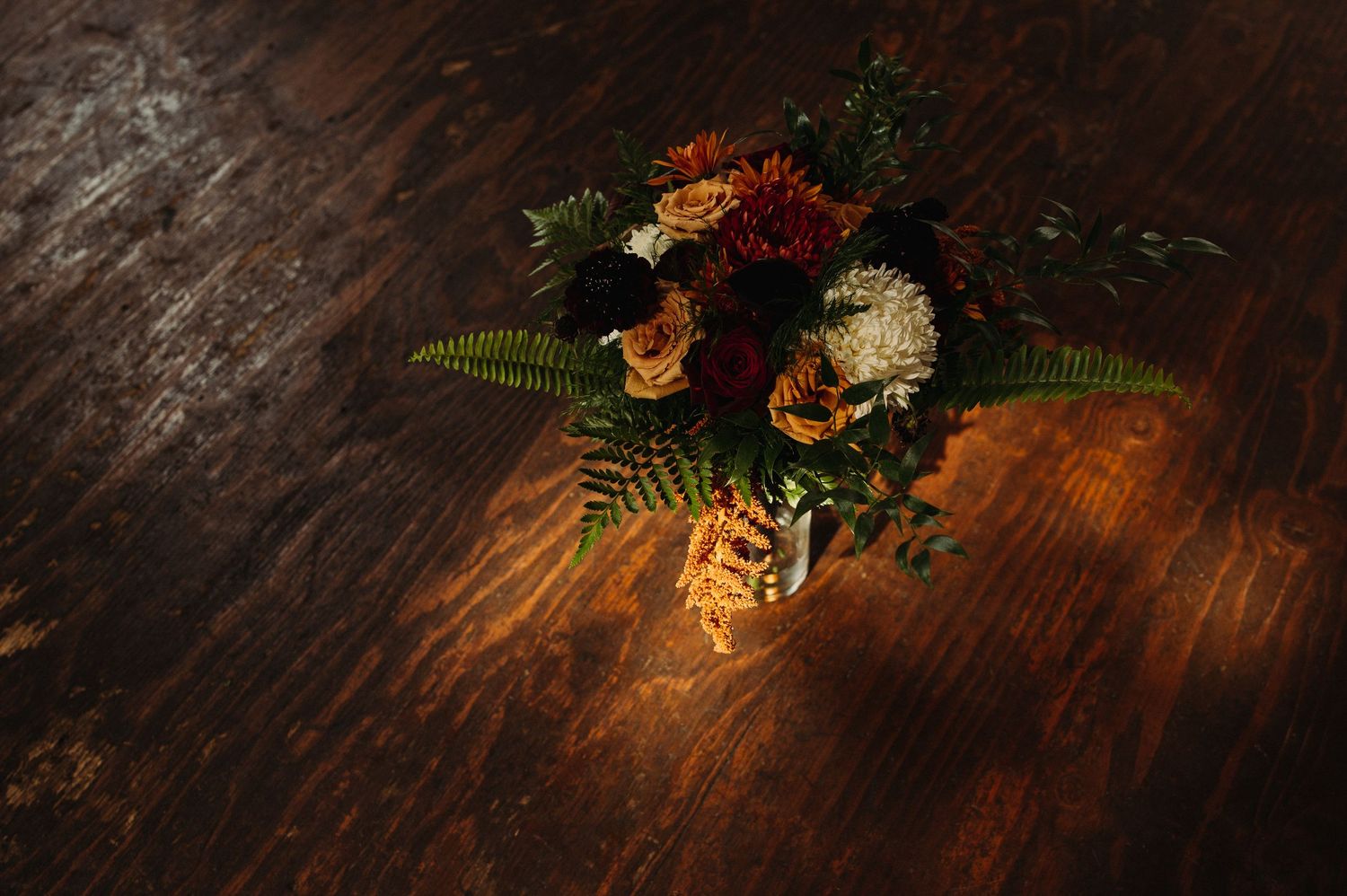 Autumn floral arrangement with ferns and orange flowers on dark wooden table in dramatic lighting.