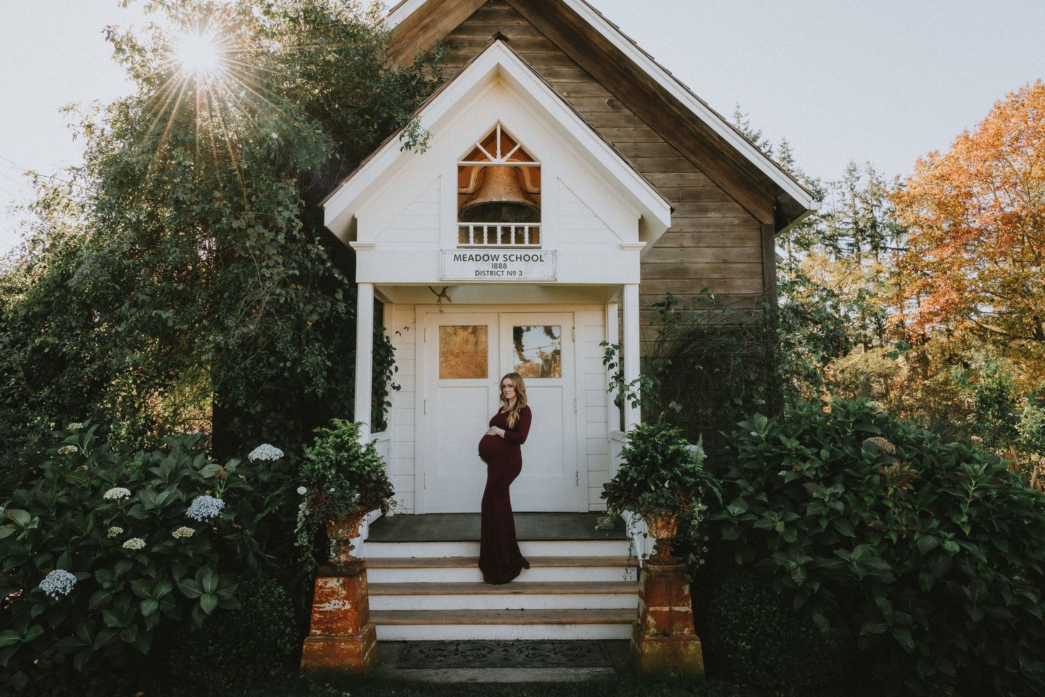 A person in a burgundy dress stands before a charming white cottage with fall foliage.