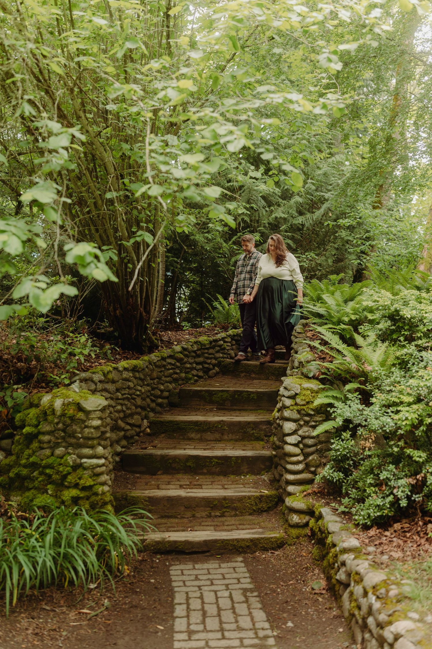 Couple walks down mossy stone steps surrounded by lush green ferns and trees in a serene garden setting.