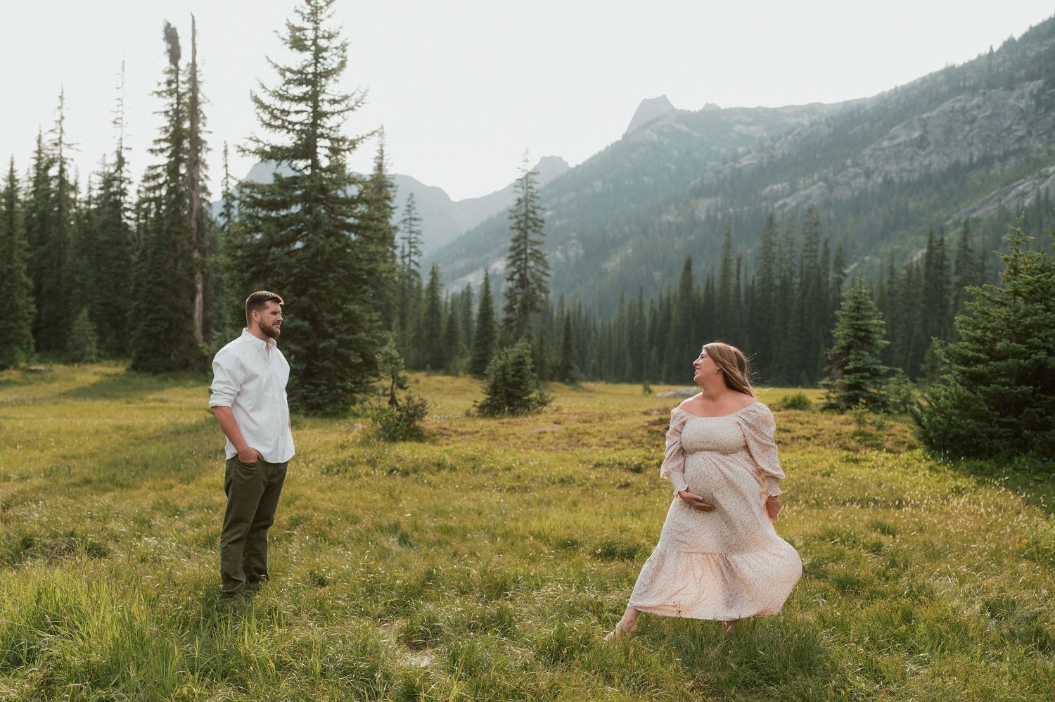 Two people share a romantic moment in an alpine meadow surrounded by evergreen trees and mountain peaks.