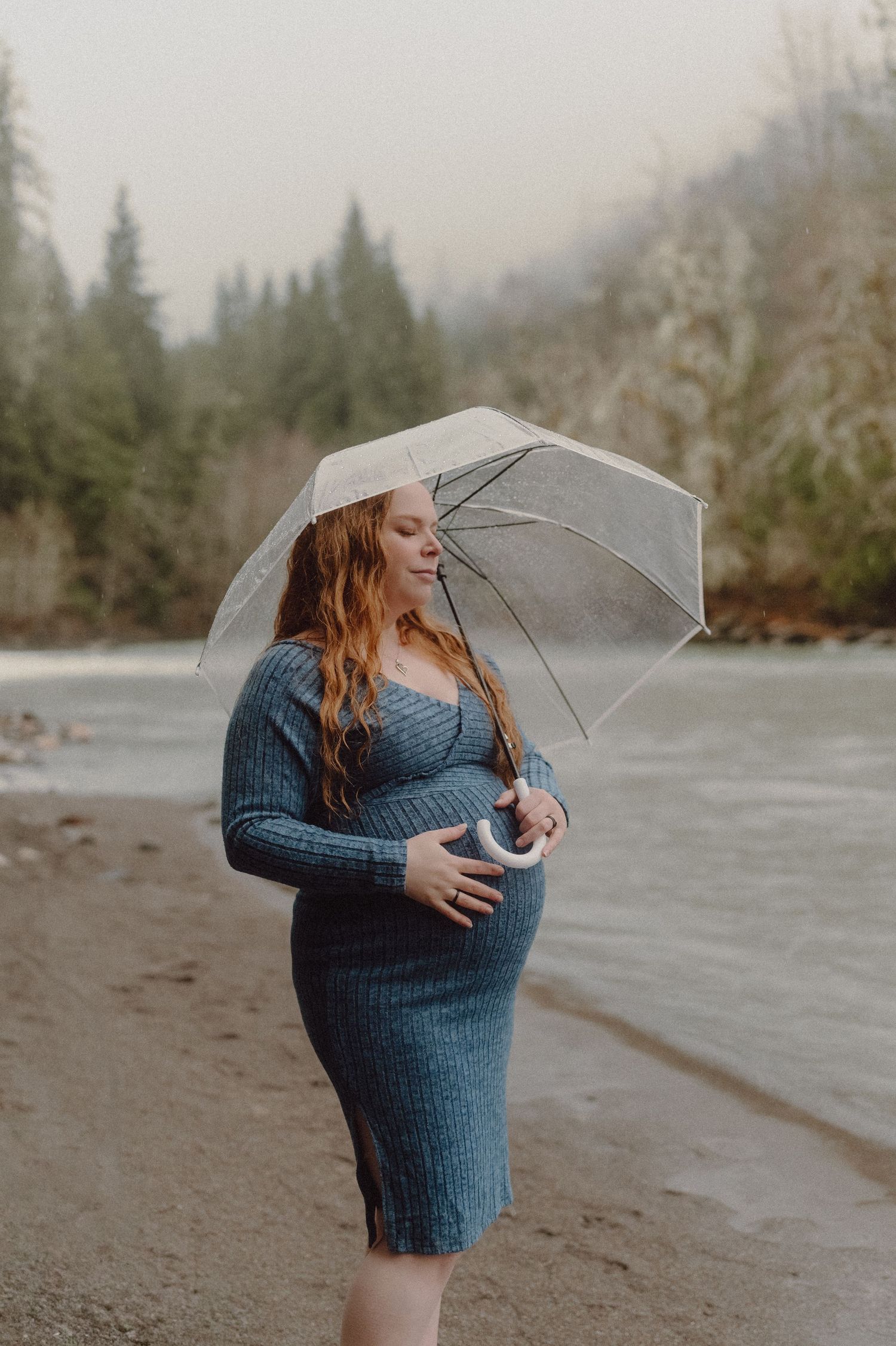 Pregnant woman in blue dress holding clear umbrella by a misty river surrounded by evergreen trees.