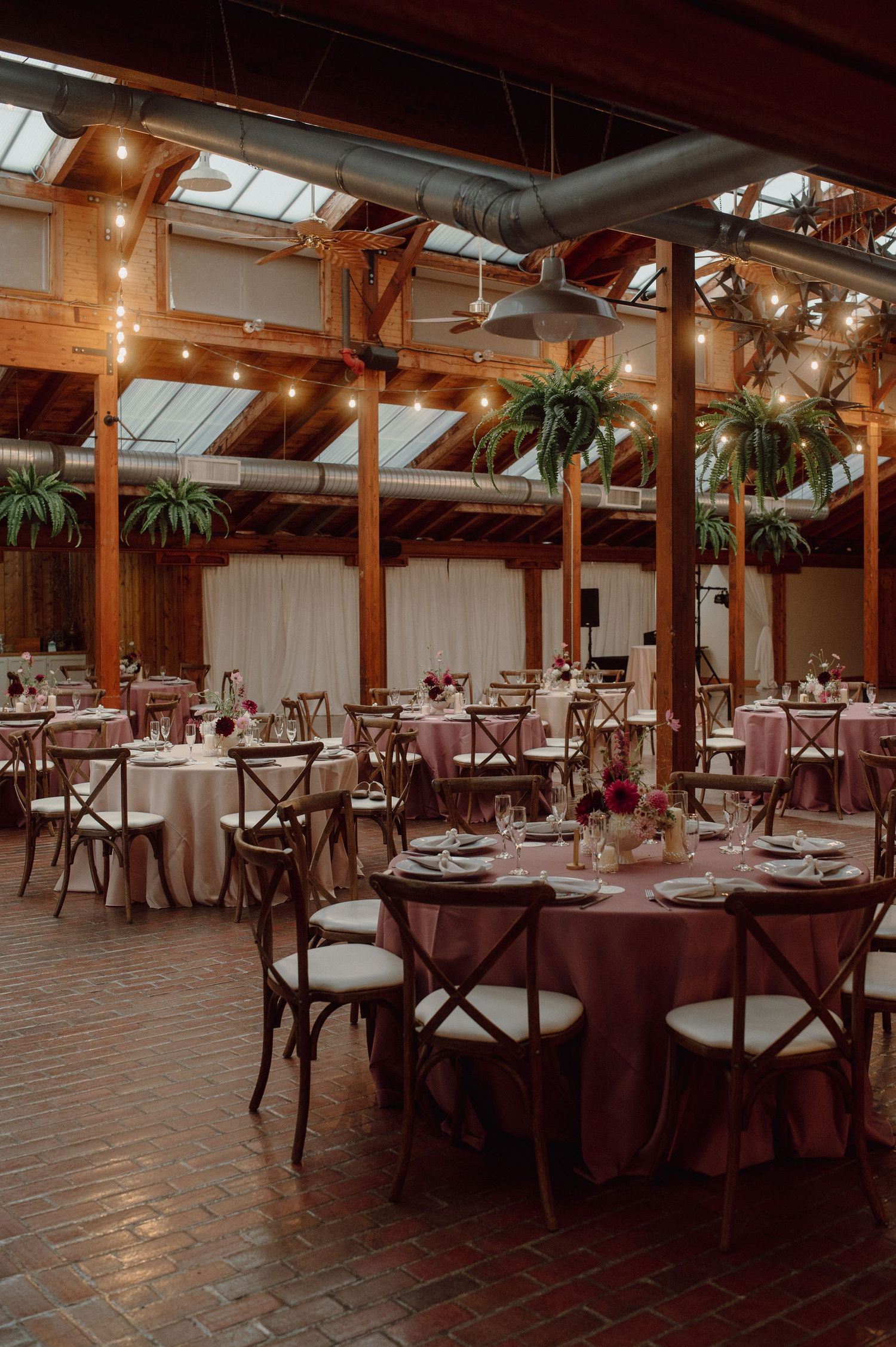 Elegant indoor wedding reception venue with wooden beams, hanging plants, and tables set for dining under skylights.