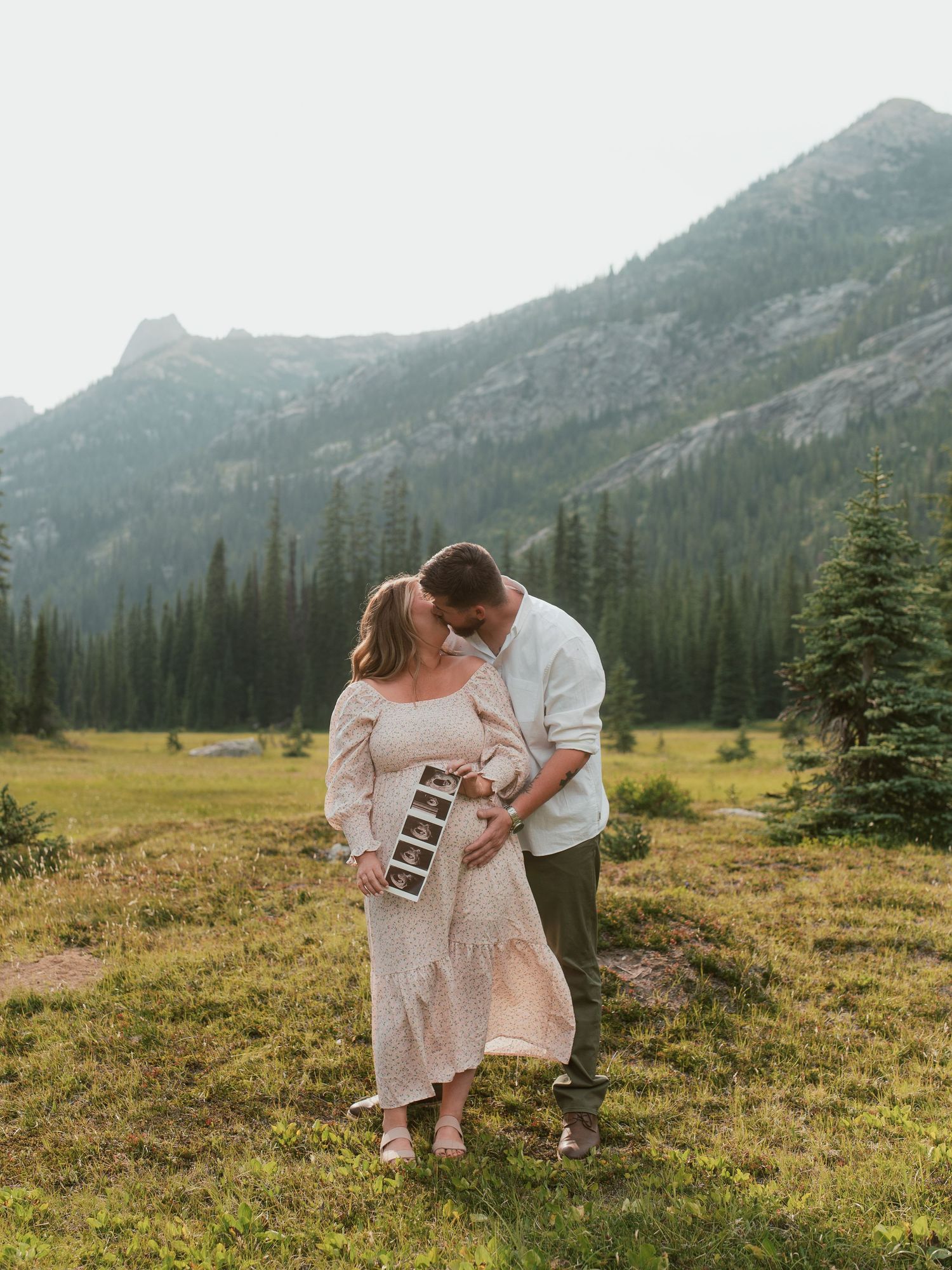 A couple shares an intimate moment in a mountain meadow with evergreen trees and peaks in the misty background.