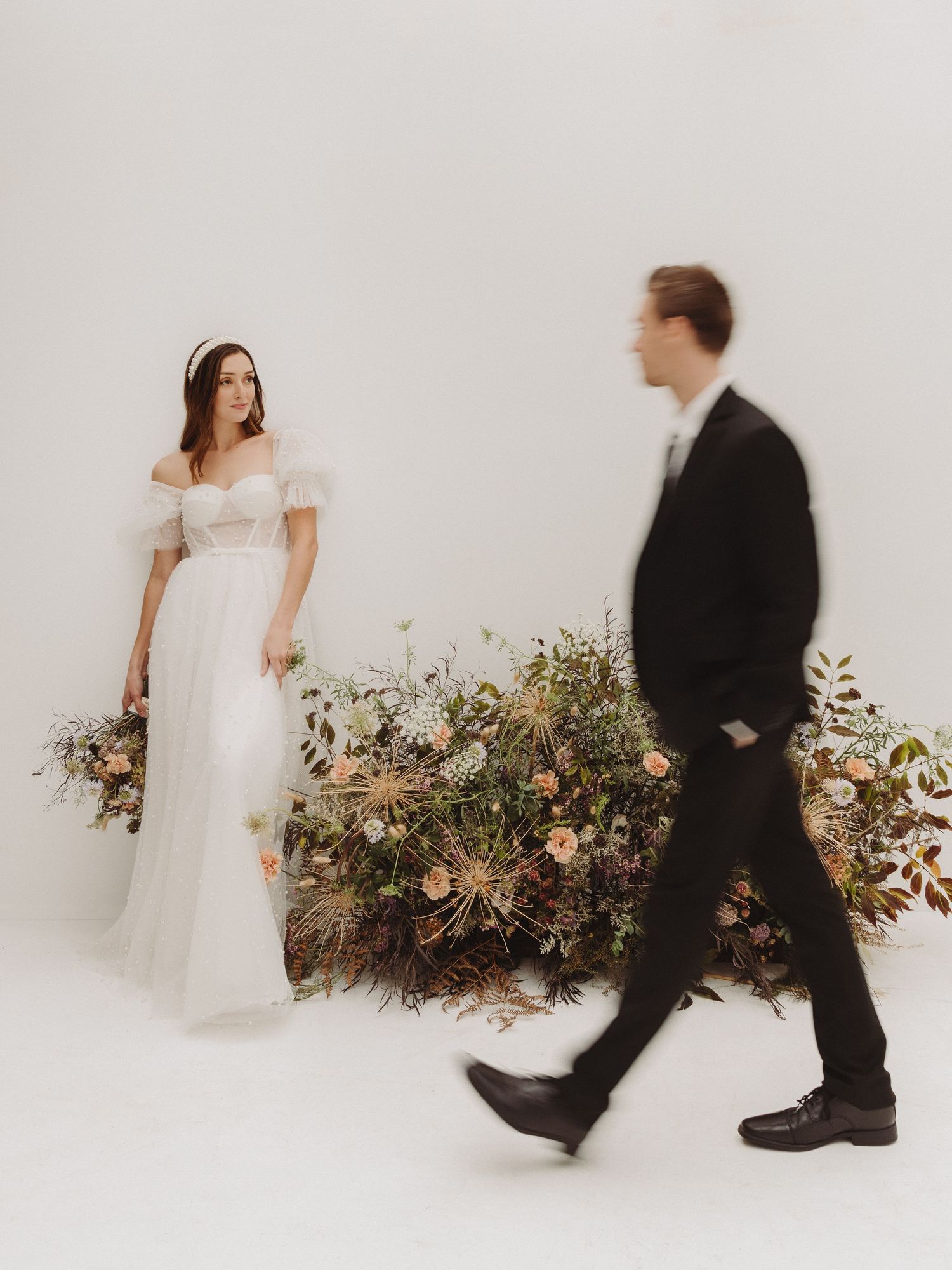 A couple in wedding attire poses against a white backdrop with floral arrangements.