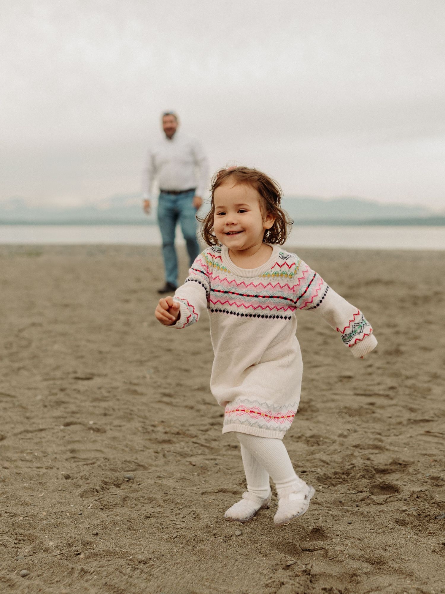 A toddler in a white patterned sweater dress runs joyfully across the sand at the beach.