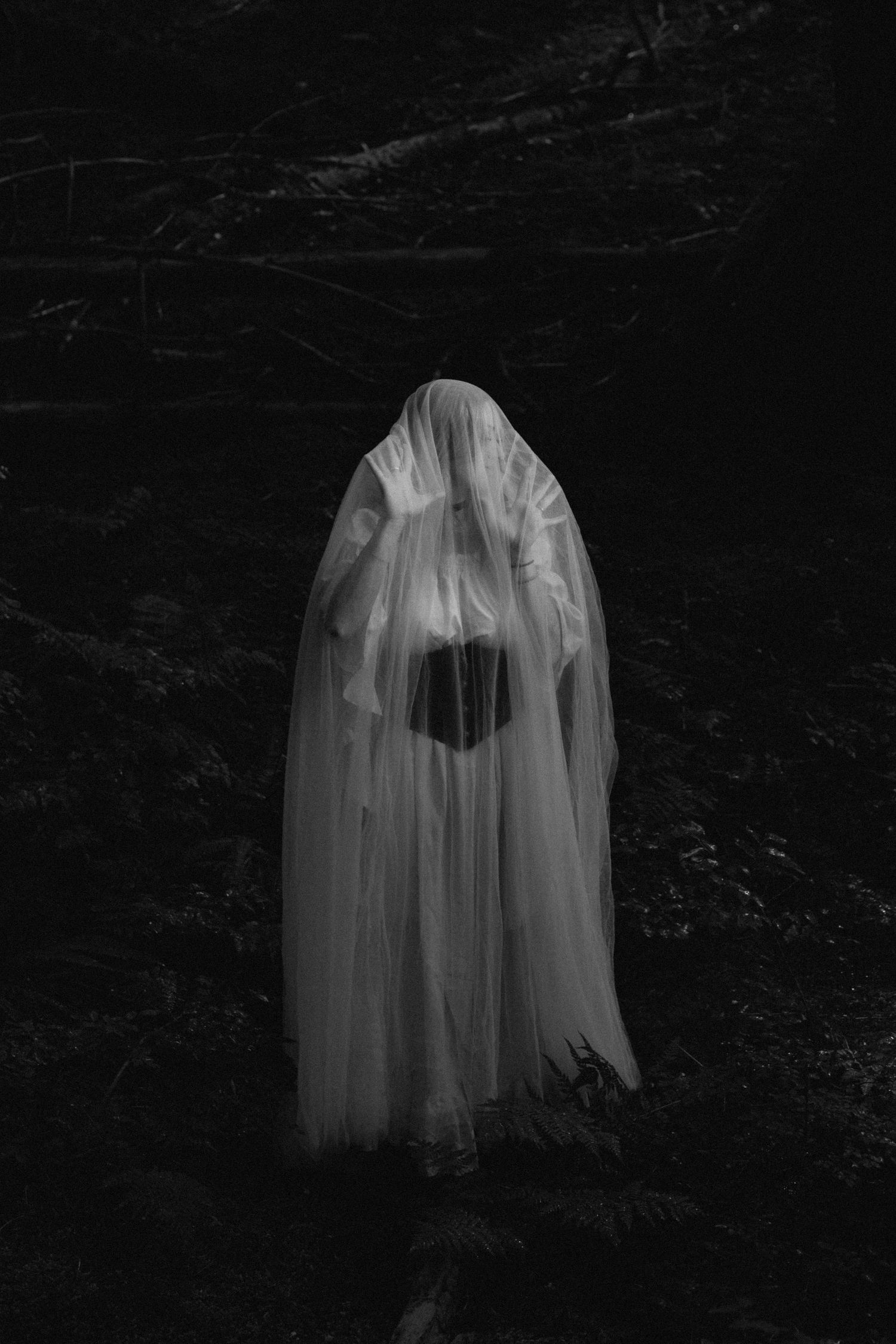 A ghostly figure in white fabric moves through a dark forest creating an eerie supernatural atmosphere in multiple exposures.