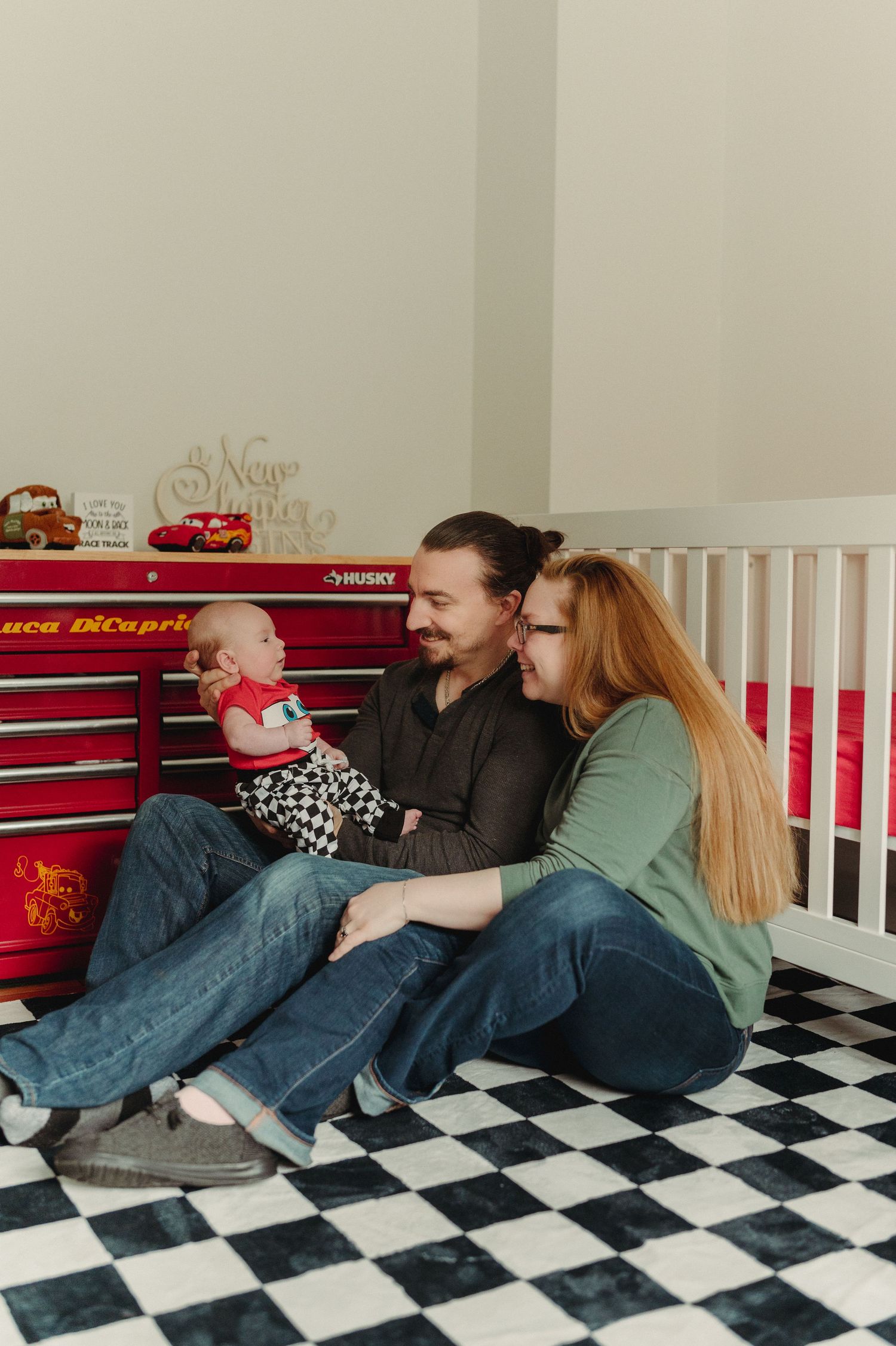 A couple sits on a checkered floor holding their baby in a Cars-themed nursery with a red Husky toolbox.